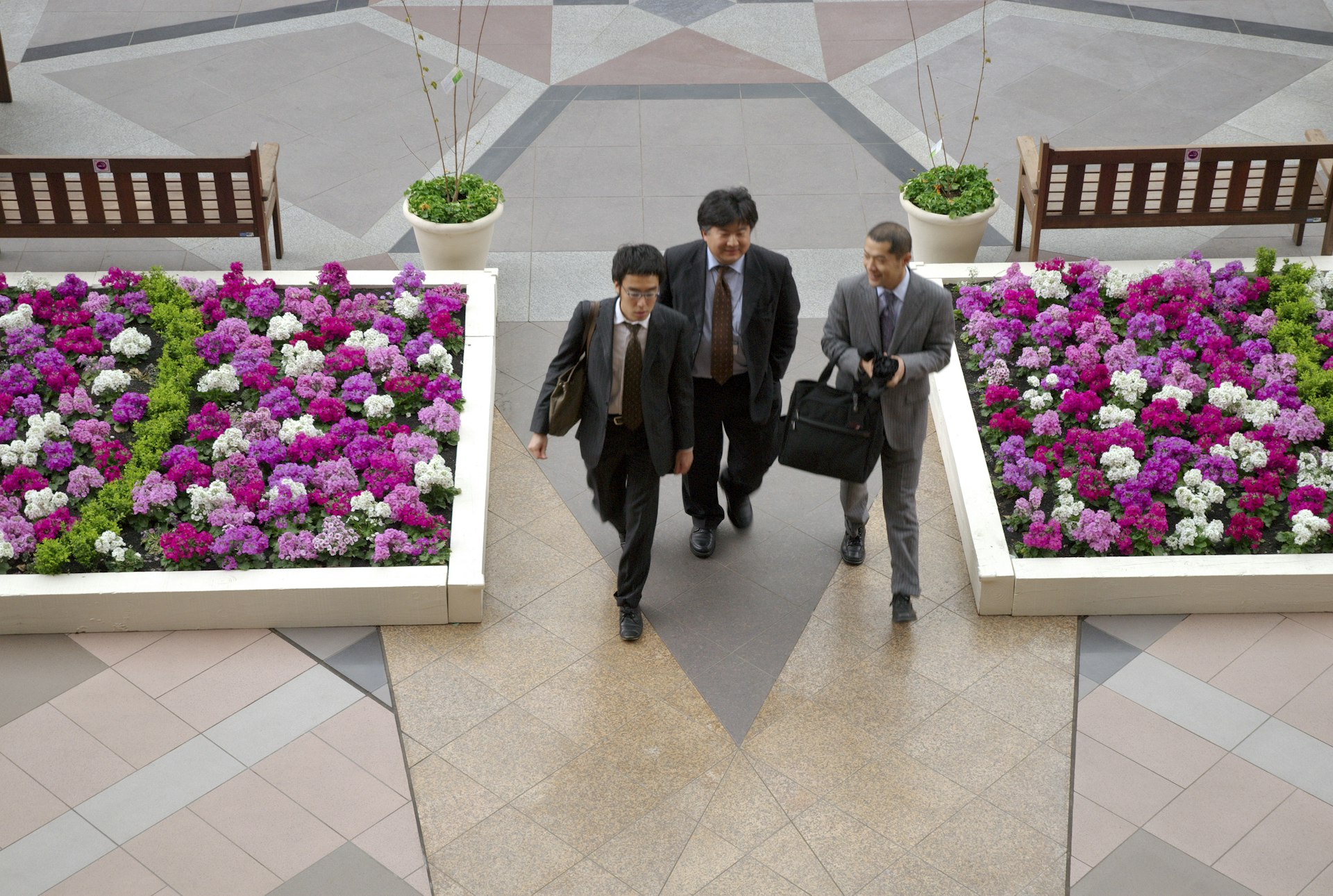 Three suited businessmen walkl past some flower beds in Yebisu Garden Place, Ebsiu in Tokyo