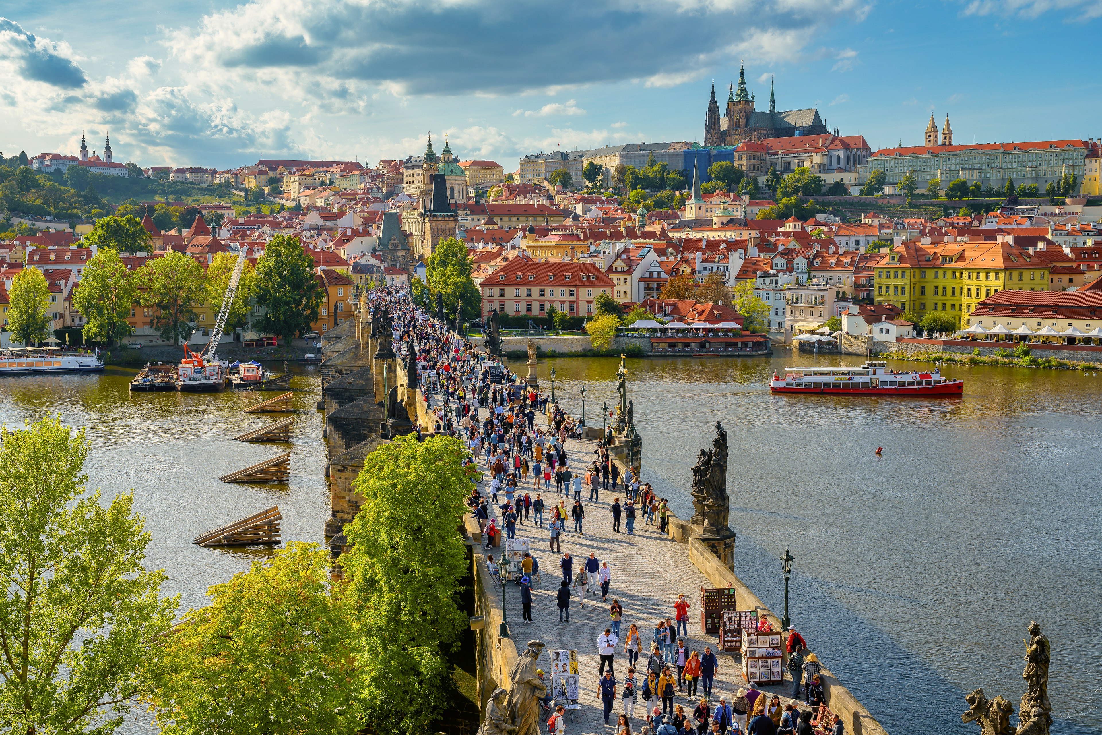 Summer tourists crowd a bridge over a river that leads to a hilltop castle on a sunny day