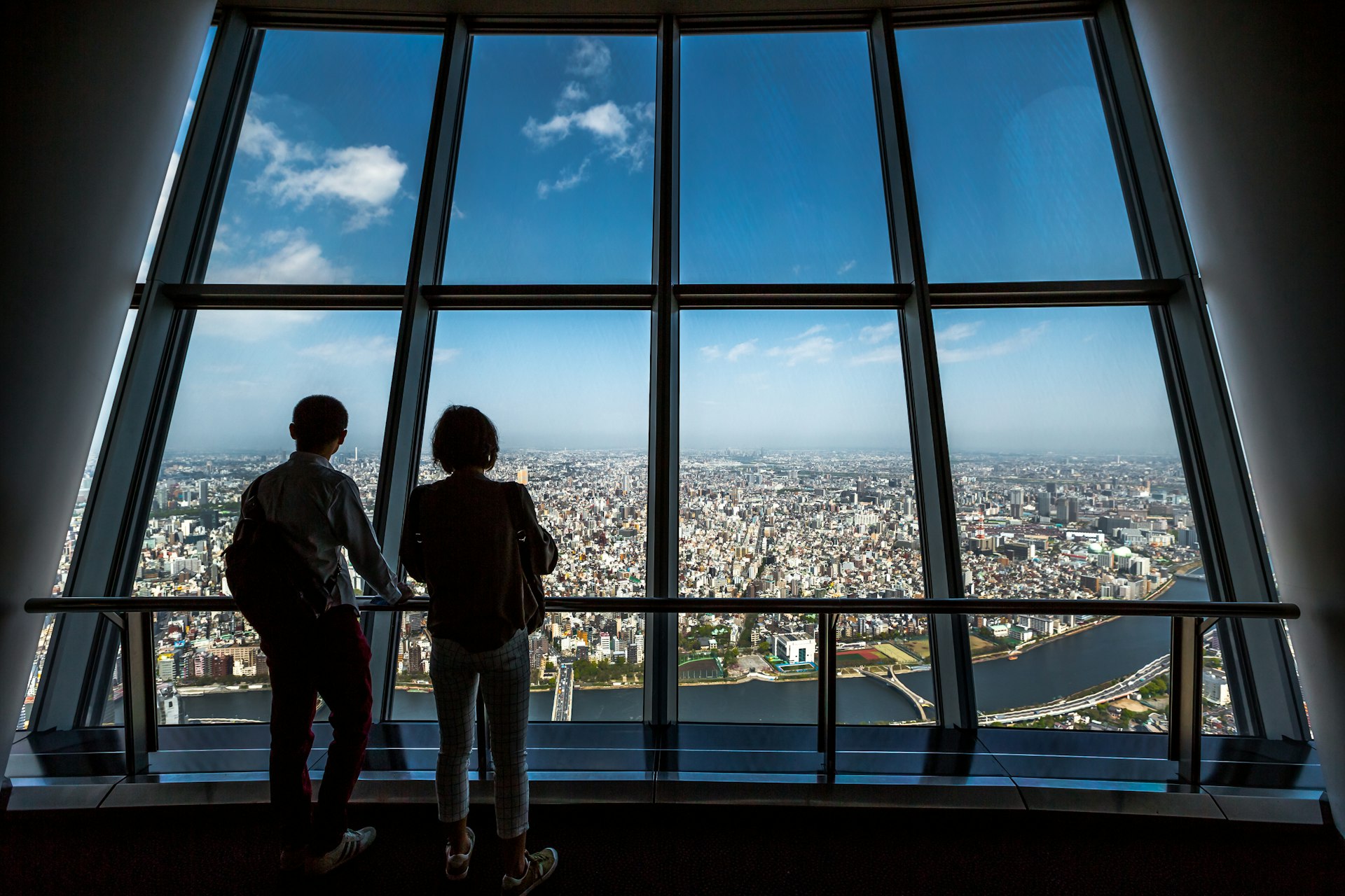 Two tourists looking out of a large window over Tokyo on the observation deck at the Tokyo Skytree