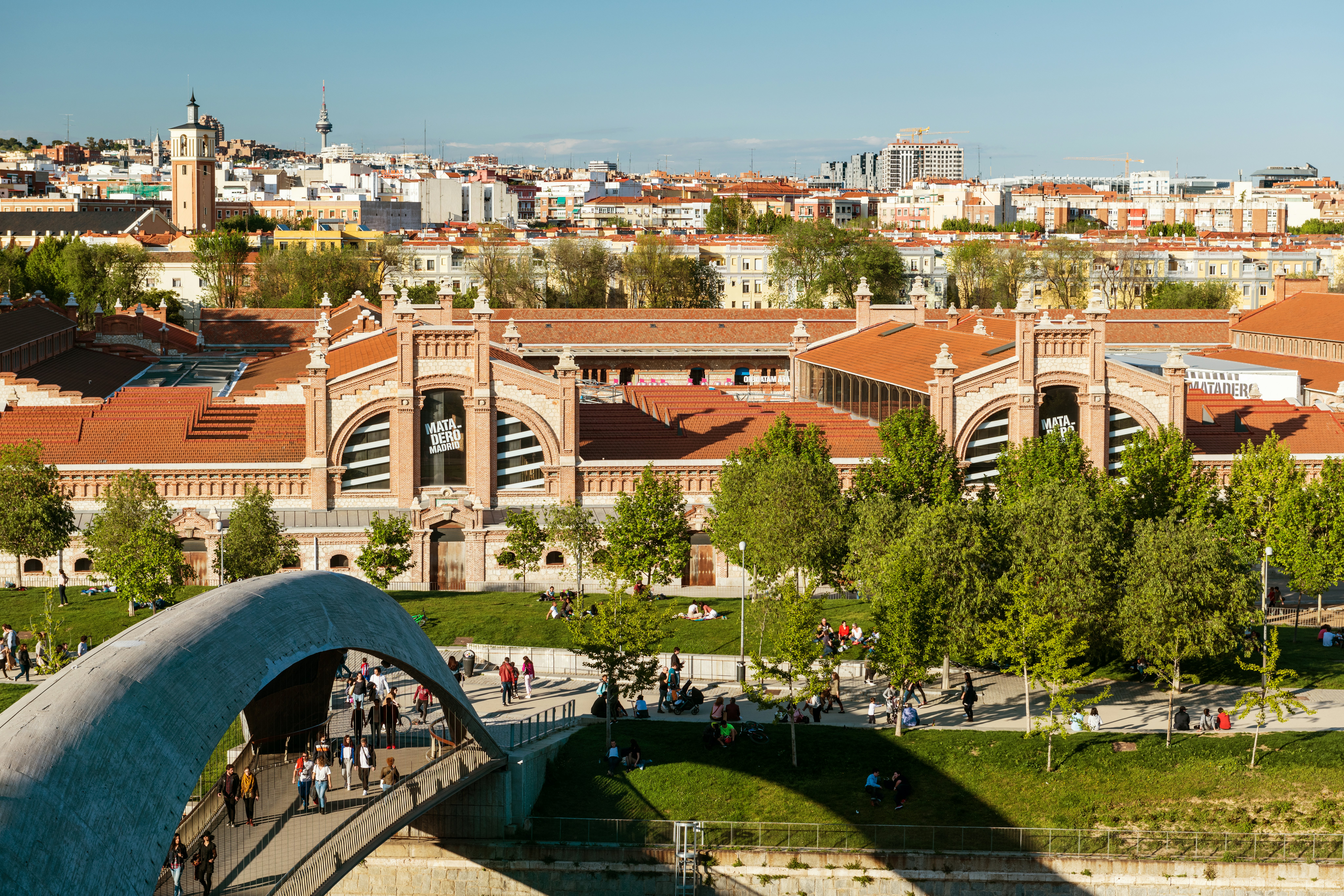 Locals and tourists walk though a modern bridge over the Manzanares River in the public park of Madrid Río in Madrid, Spain, with the Matadero building (a former slaughterhouse converted to an arts centre) in the background.
