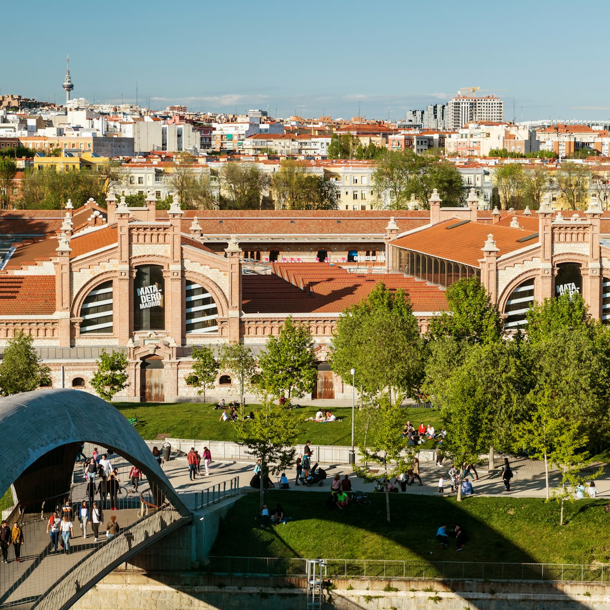 Locals and tourists walk though a modern bridge over the Manzanares River in the public park of Madrid Río in Madrid, Spain, with the Matadero building (a former slaughterhouse converted to an arts centre) in the background.