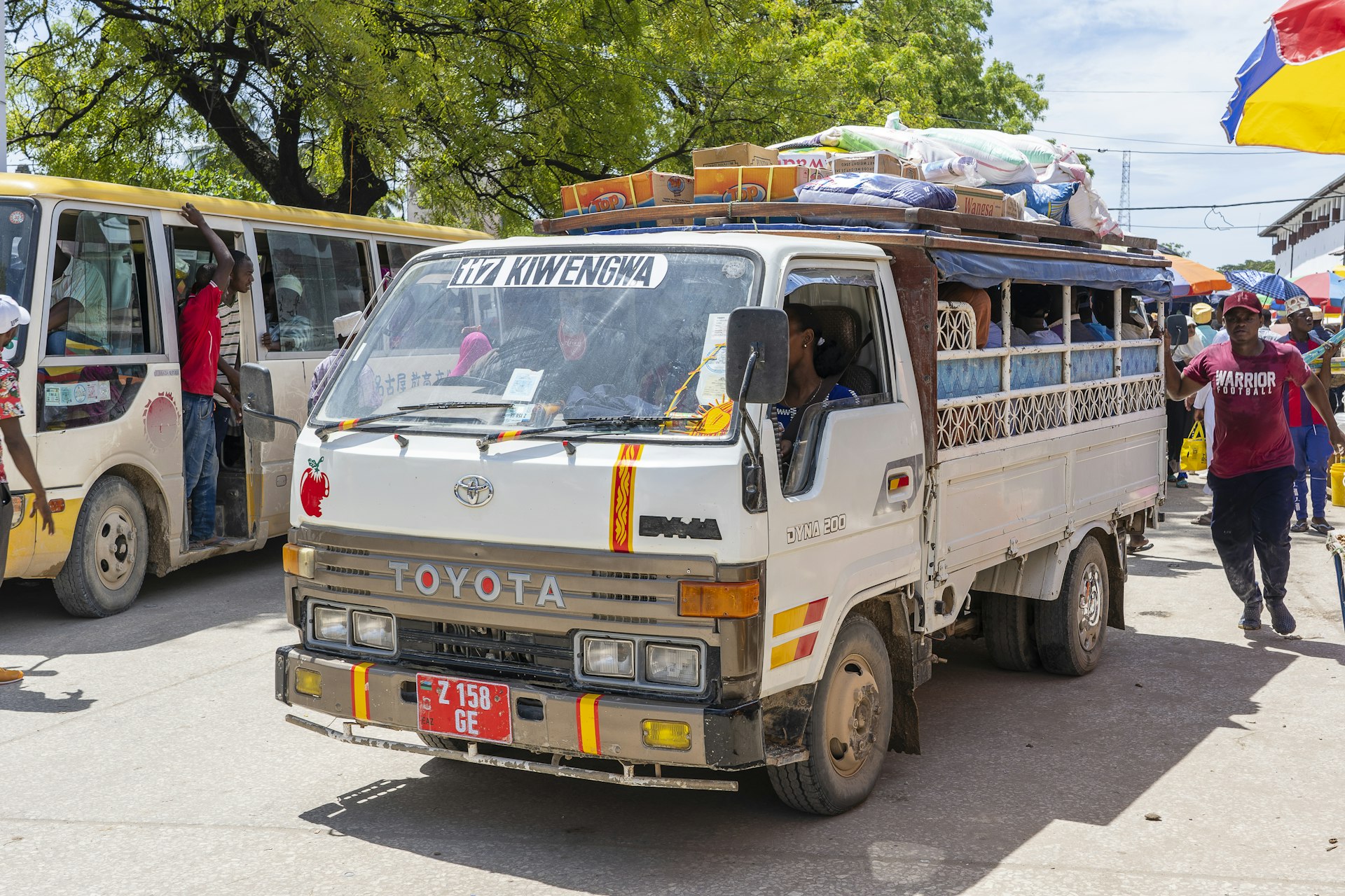 A Dala dala bus, on the streets of Tanzania, alongside more public transport, with passengers and pedestrians in the background