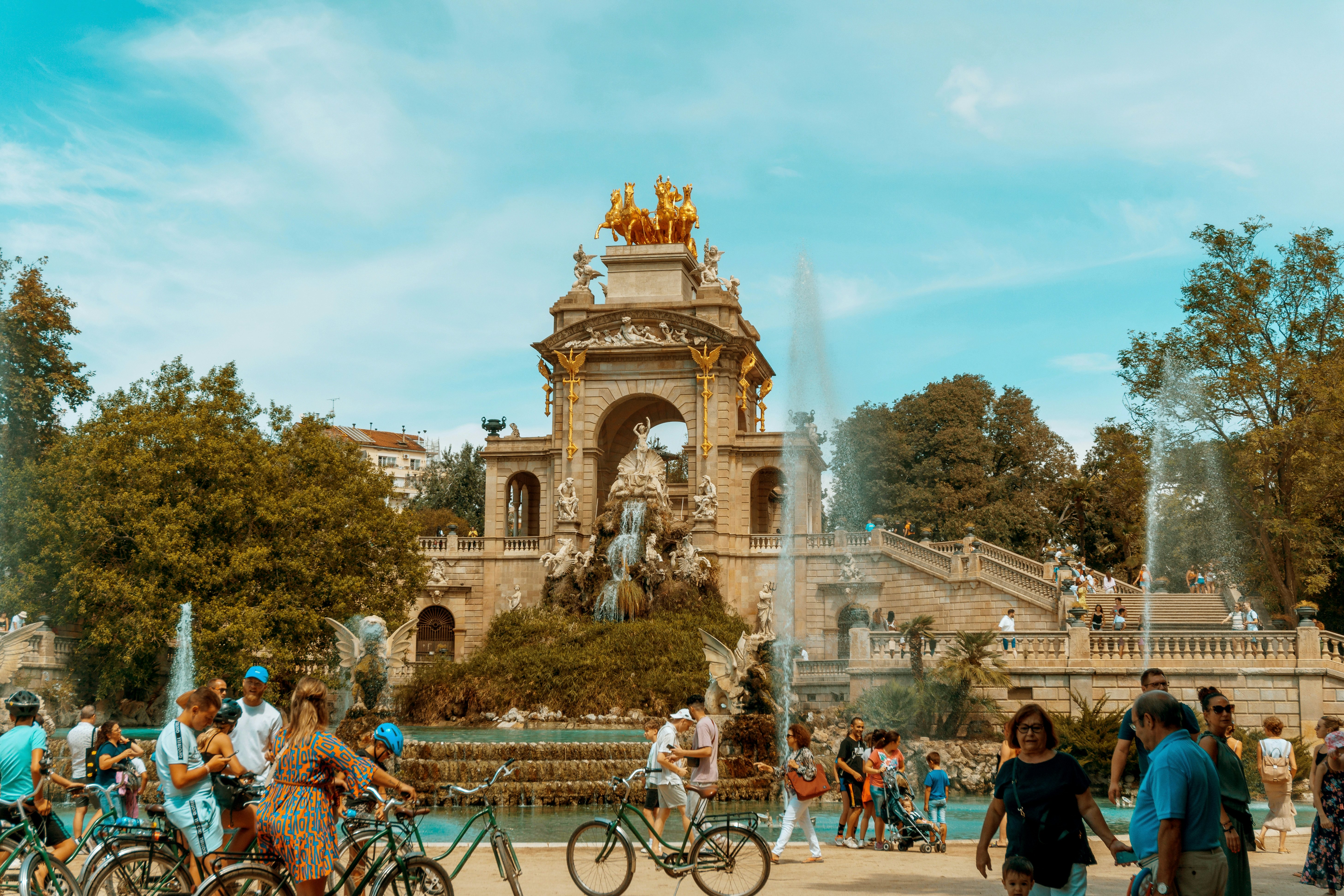 People walking and biking in front of an elaborate fountain in a park in Barcelona.