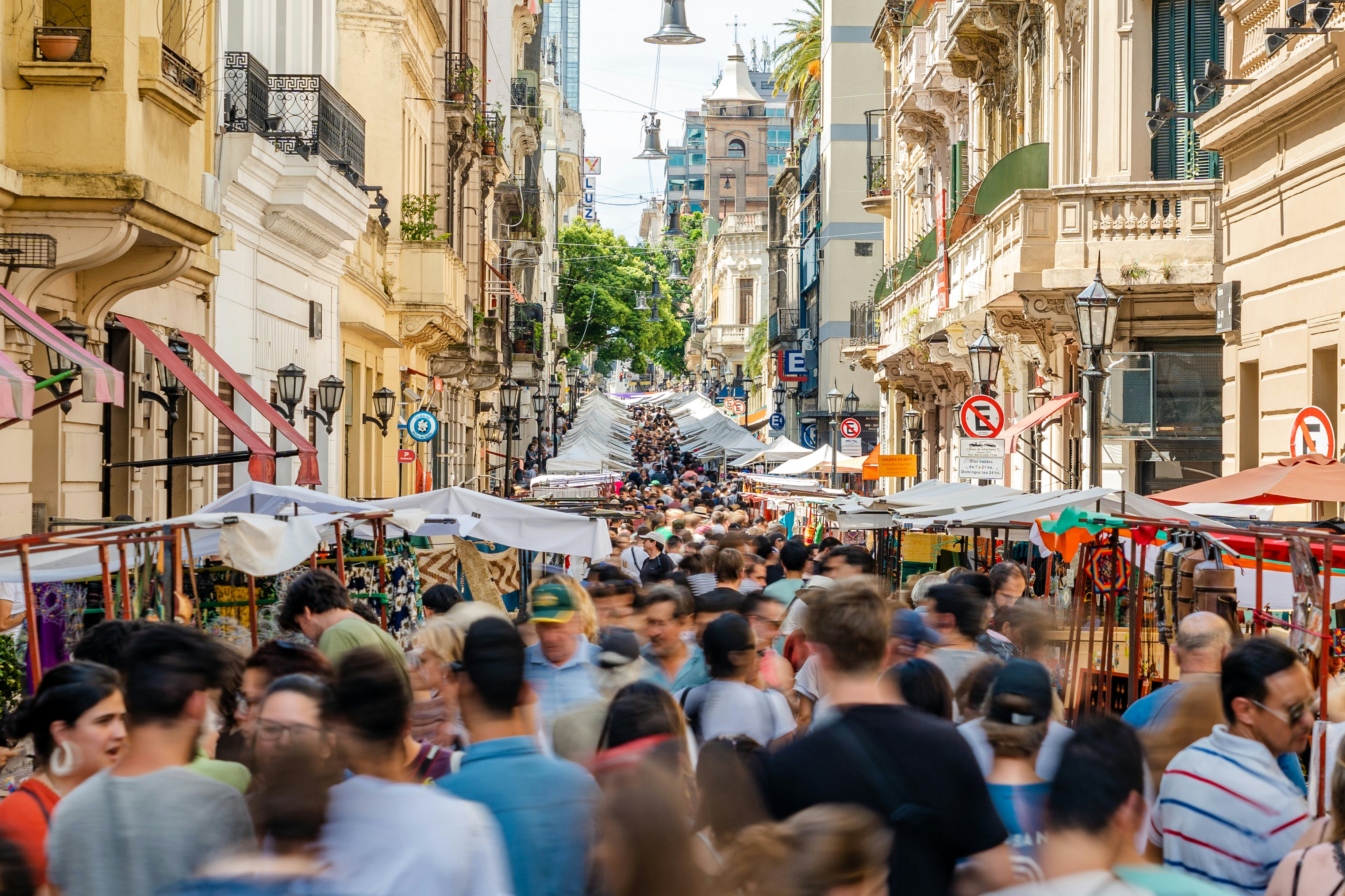 An open market takes place on a city street. The market goes on for several blocks, and the street is closed to cars and crowded with shoppers, vendors and pedestrians.