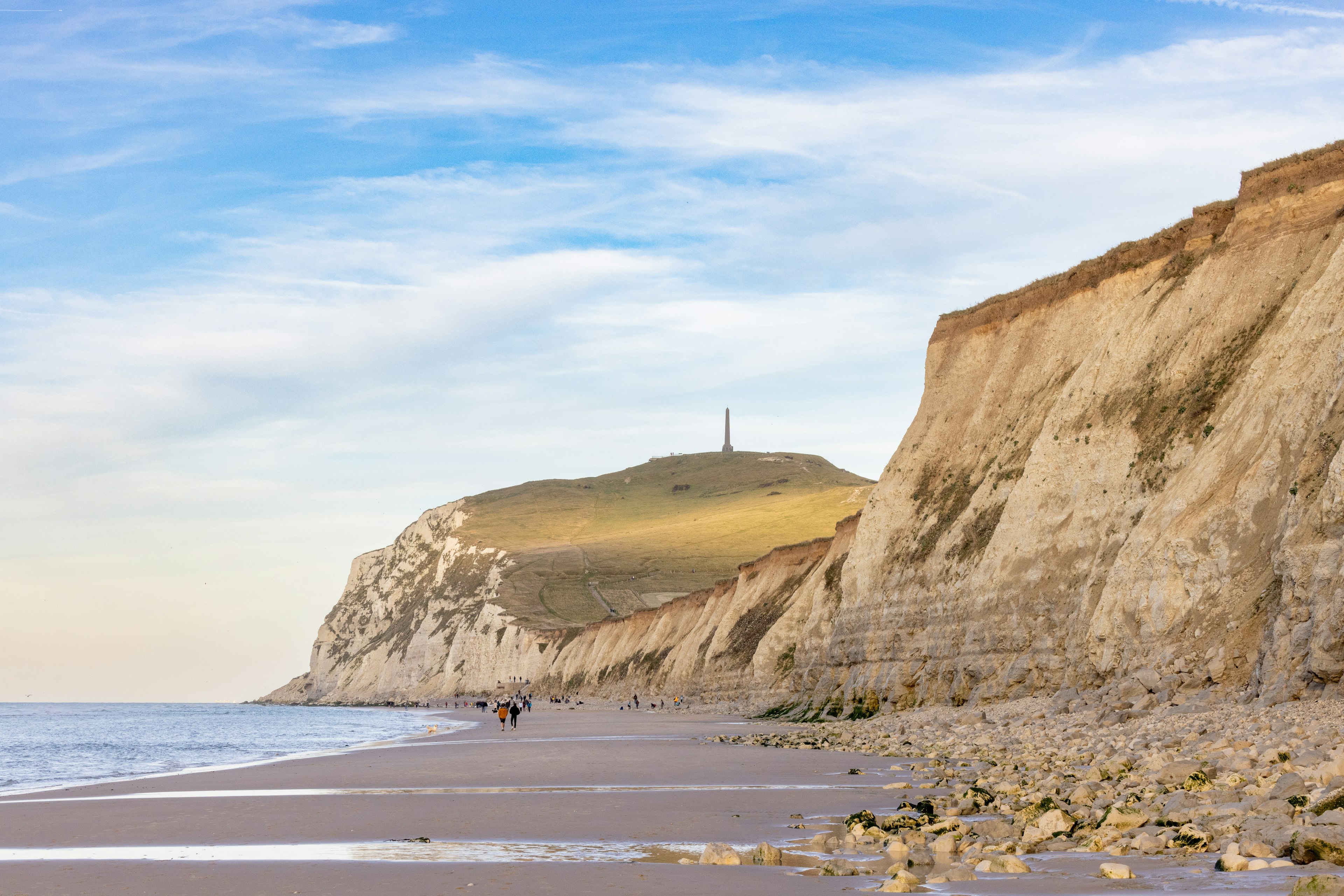 People walk along a beach beside a large cliff with an obelisk on the top