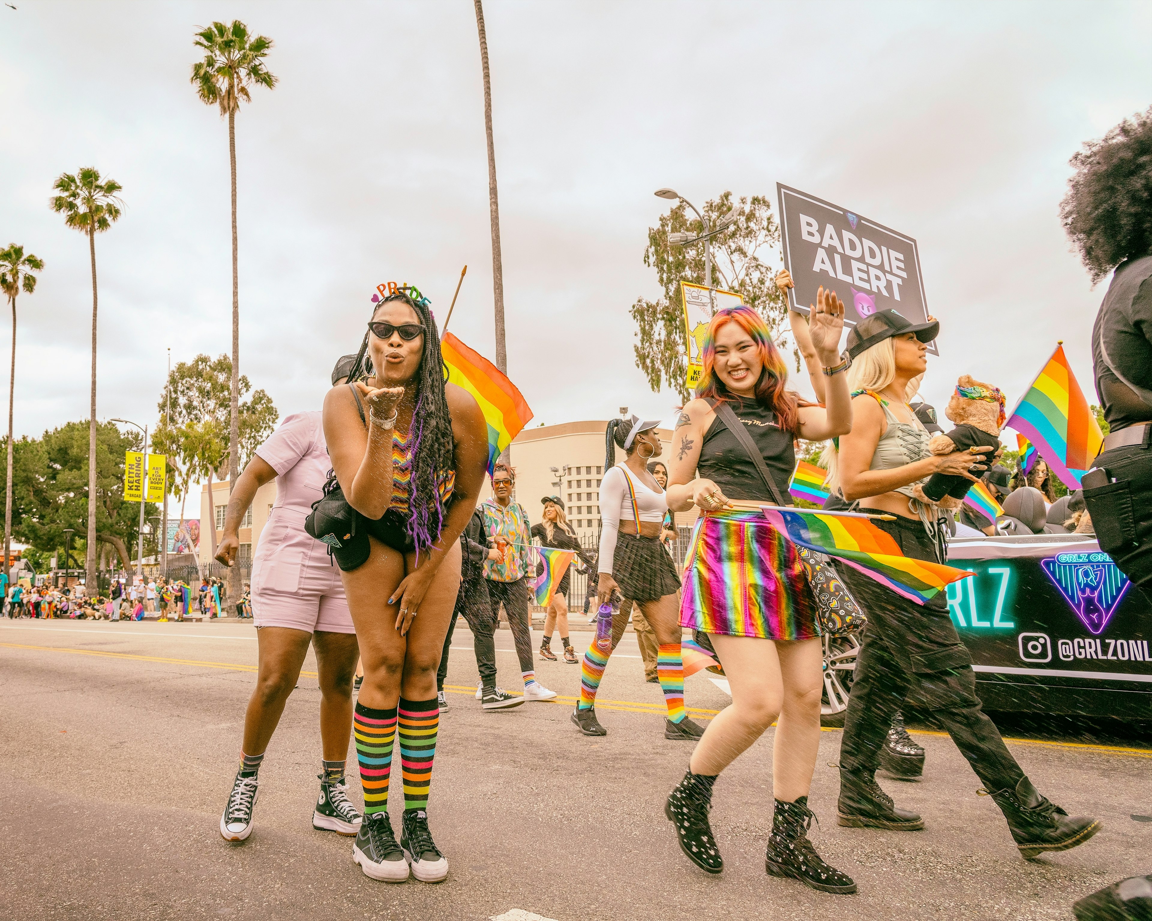 Two people dressed in rainbow colors pause as they walk in a parade to smile and blow a kiss at the camera