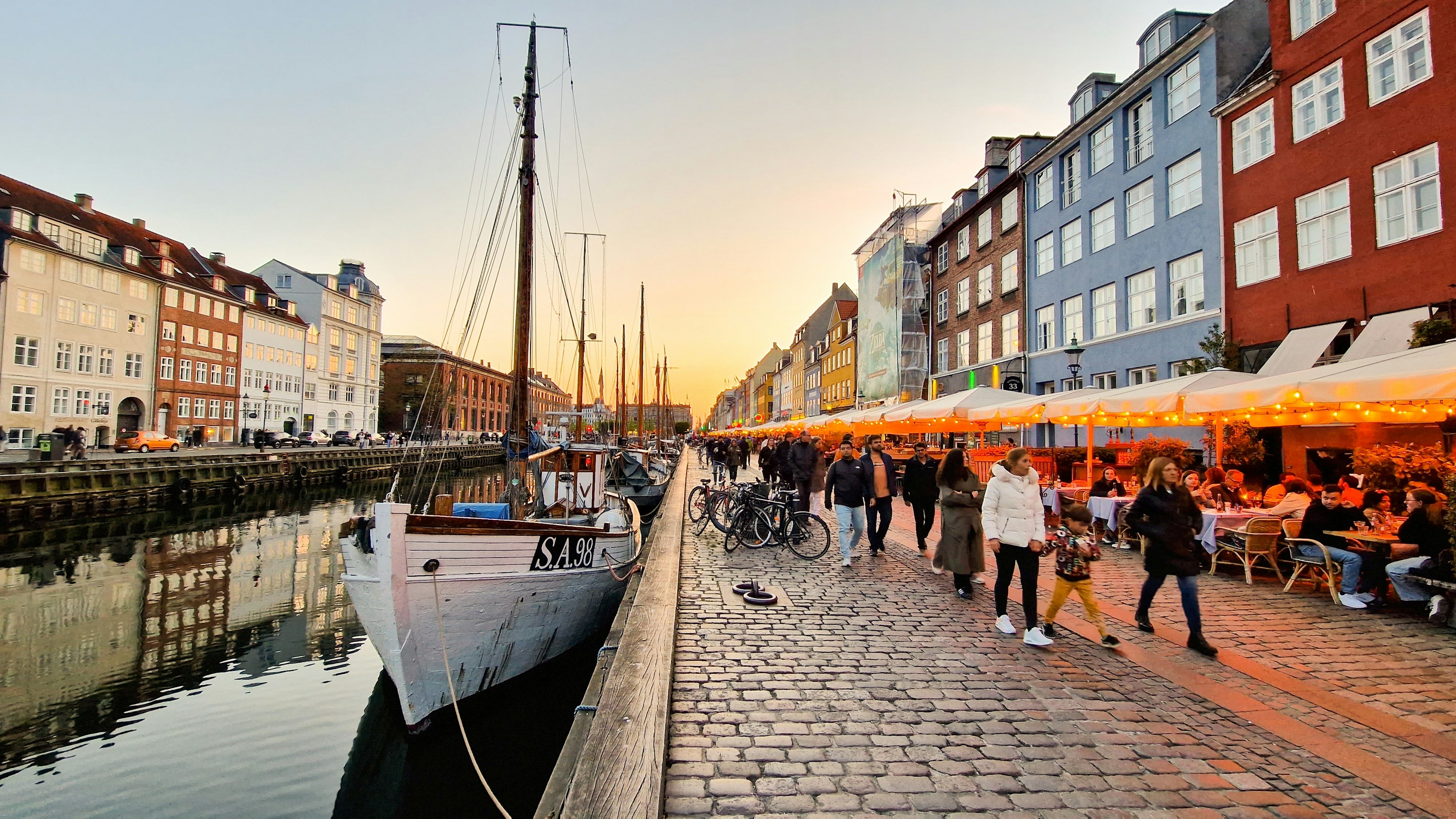 People walk by boats and past buildings at sunset in Nyhavn, Copenhagen, Denmark