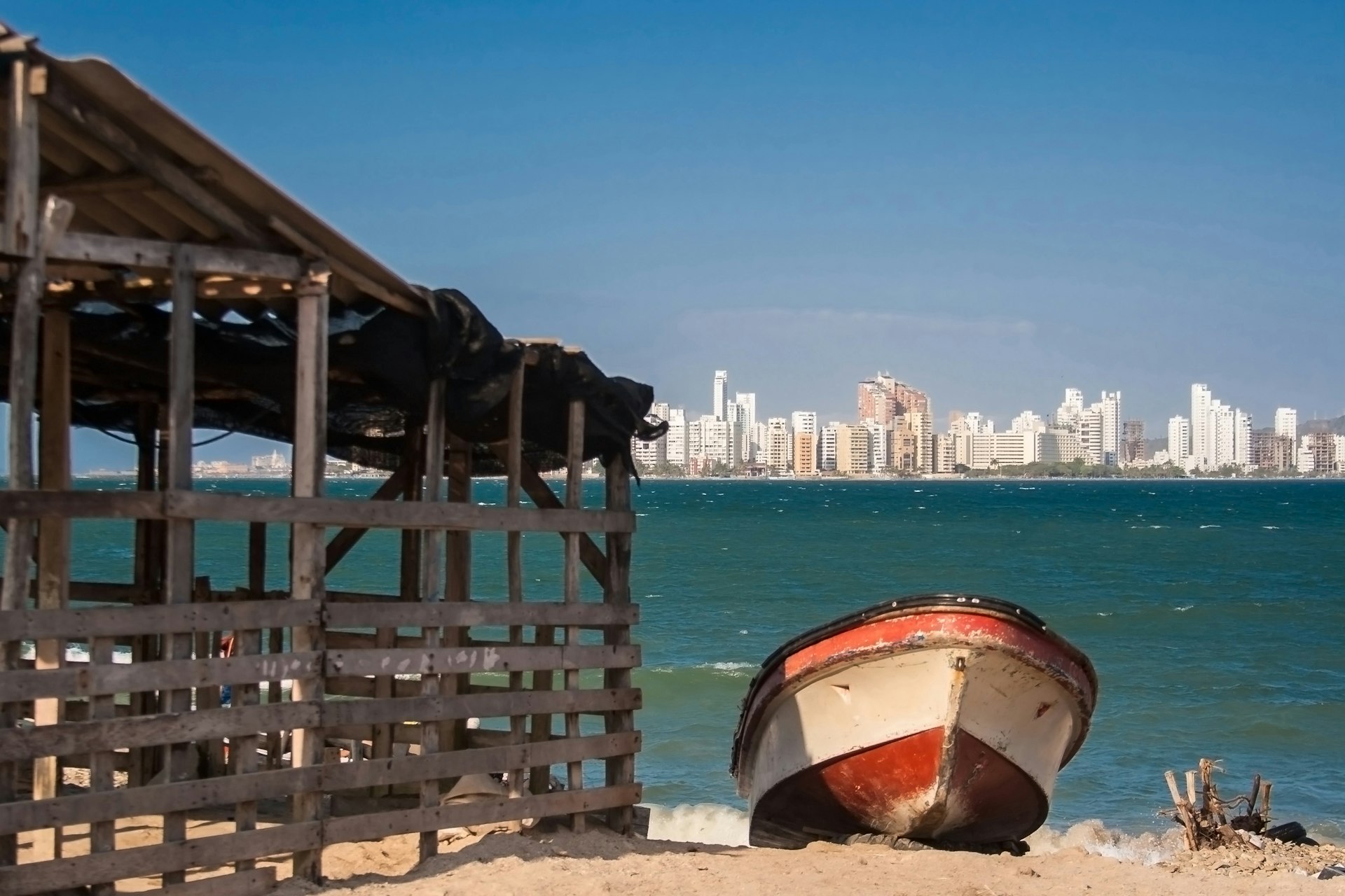 A boat rests on a sandy beach. A city skyline with high-rise buildings is in the distance across the water