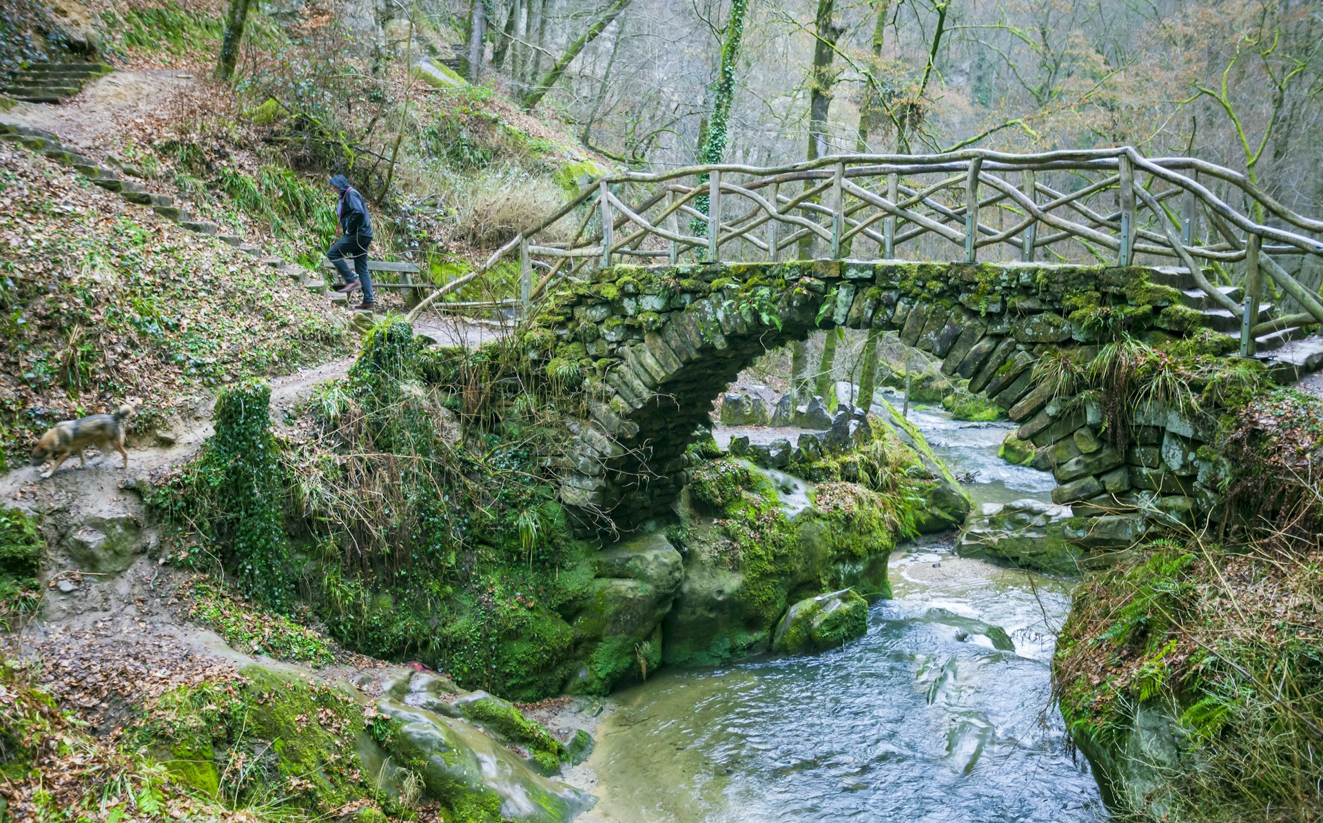 A small arched stone bridge crosses a stream with a hiker following a path through woodland