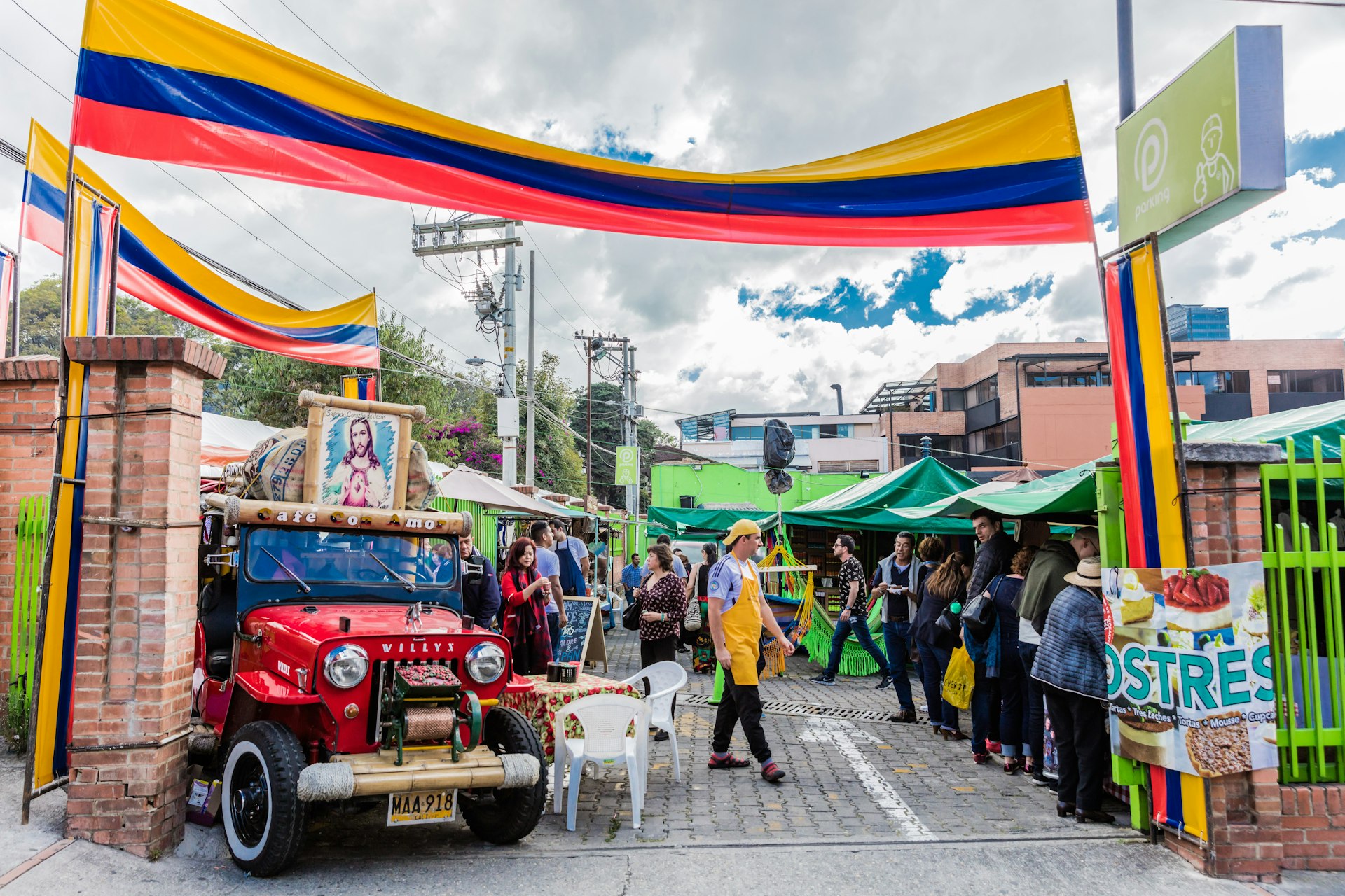 The entrance to a market flanked by stalls, a large truck, and lots of people with a red, yellow and blue Colombian flag flying overhead