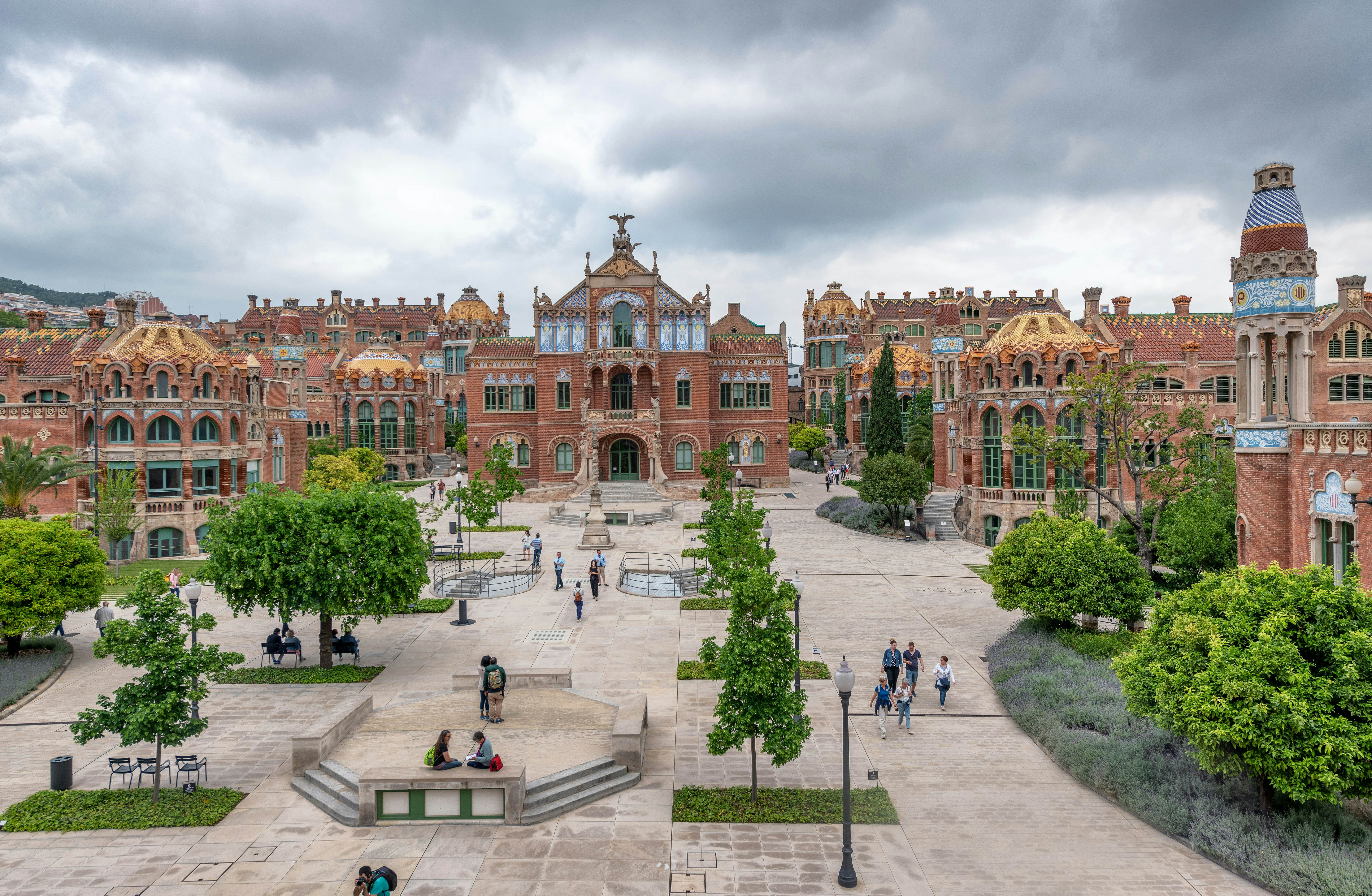 A plaza in Barcelona, Spain, has benches and green trees; it is surrounded by low redbrick buildings with exterior flourishes.