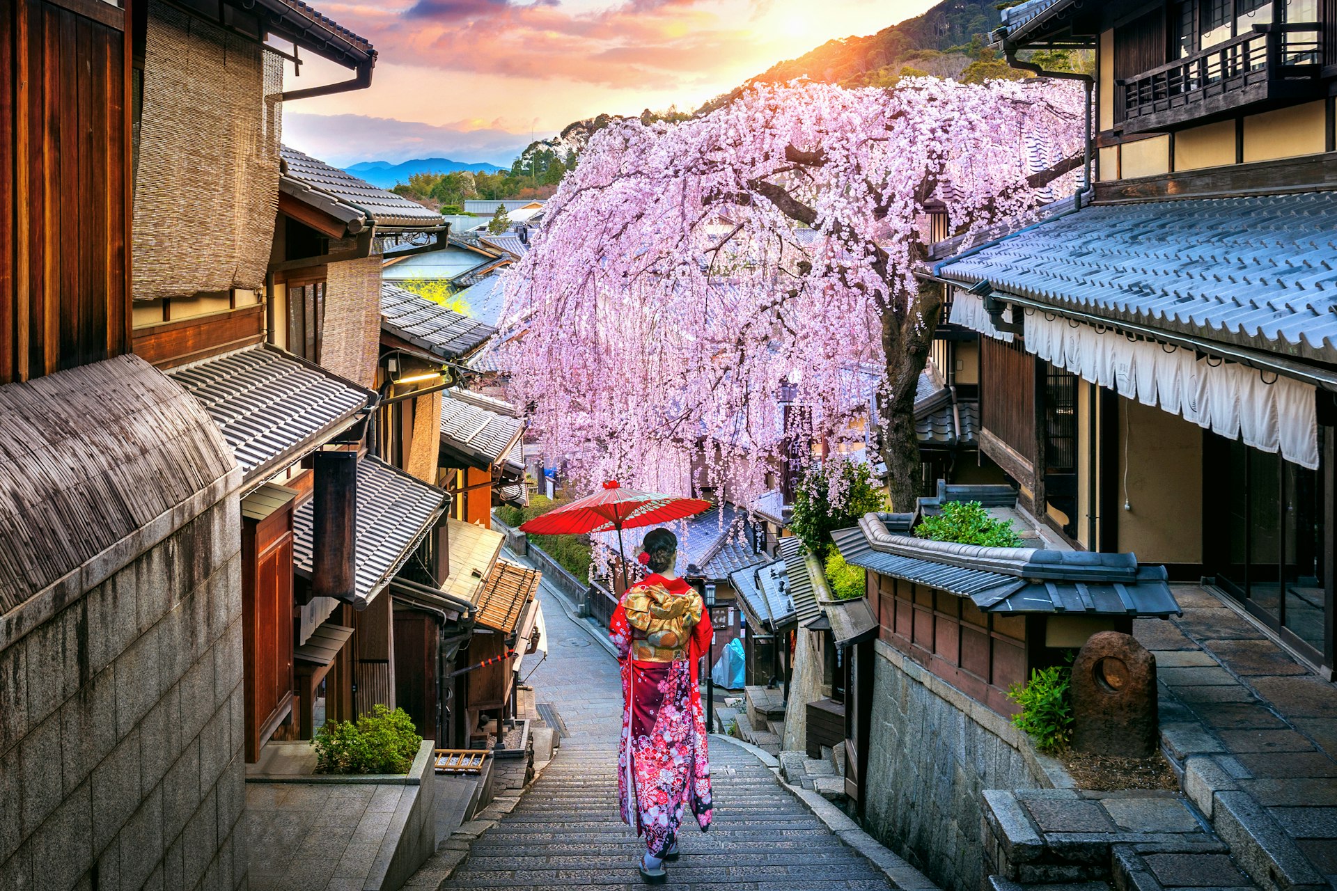 A woman in traditional Japanese dress carrying a painted umbrella walks down a path between historic wooden houses