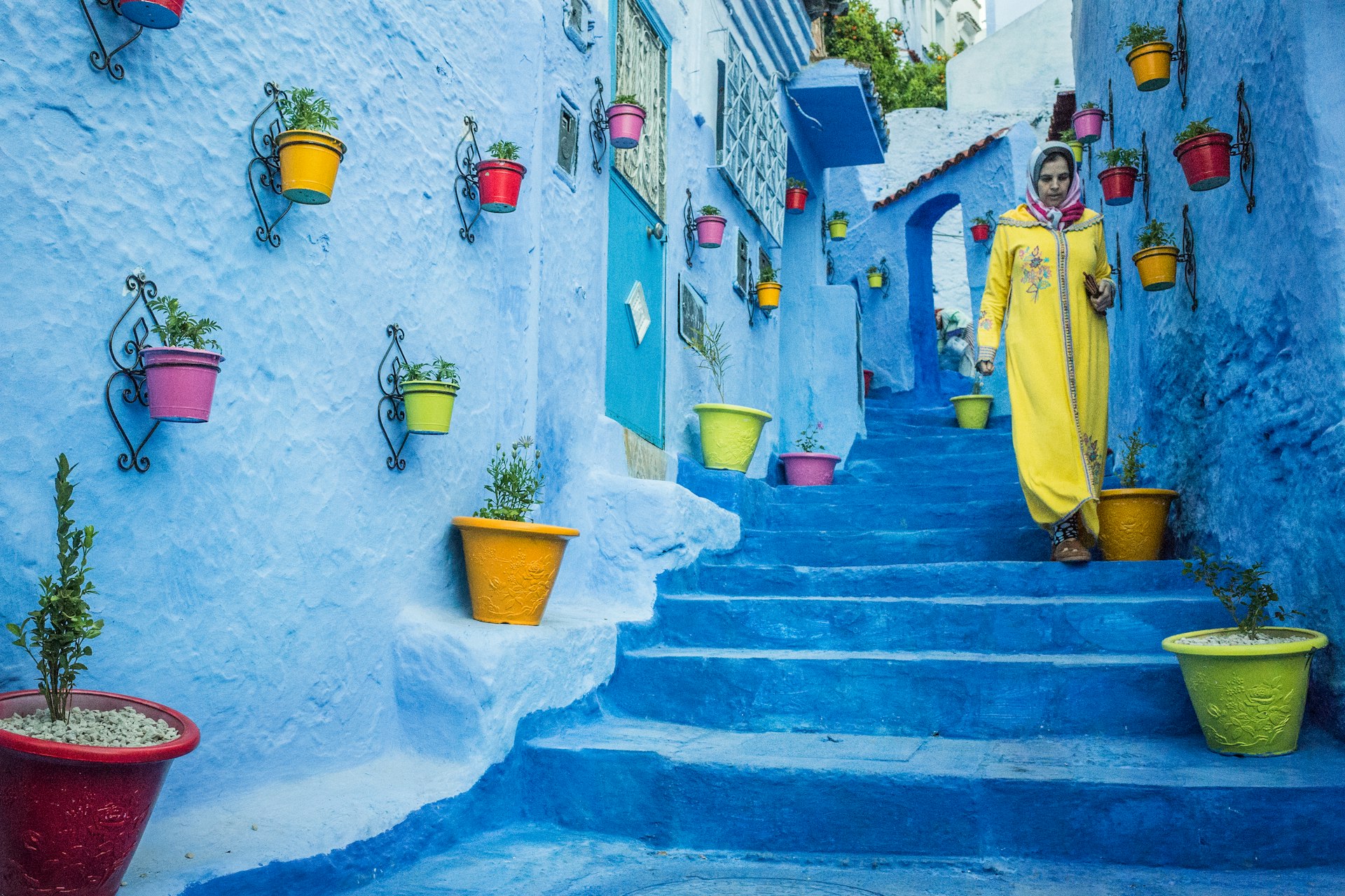 A woman in a yellow abaya walks by blue steps and walls on a street in Chefchaouen, Morocco
