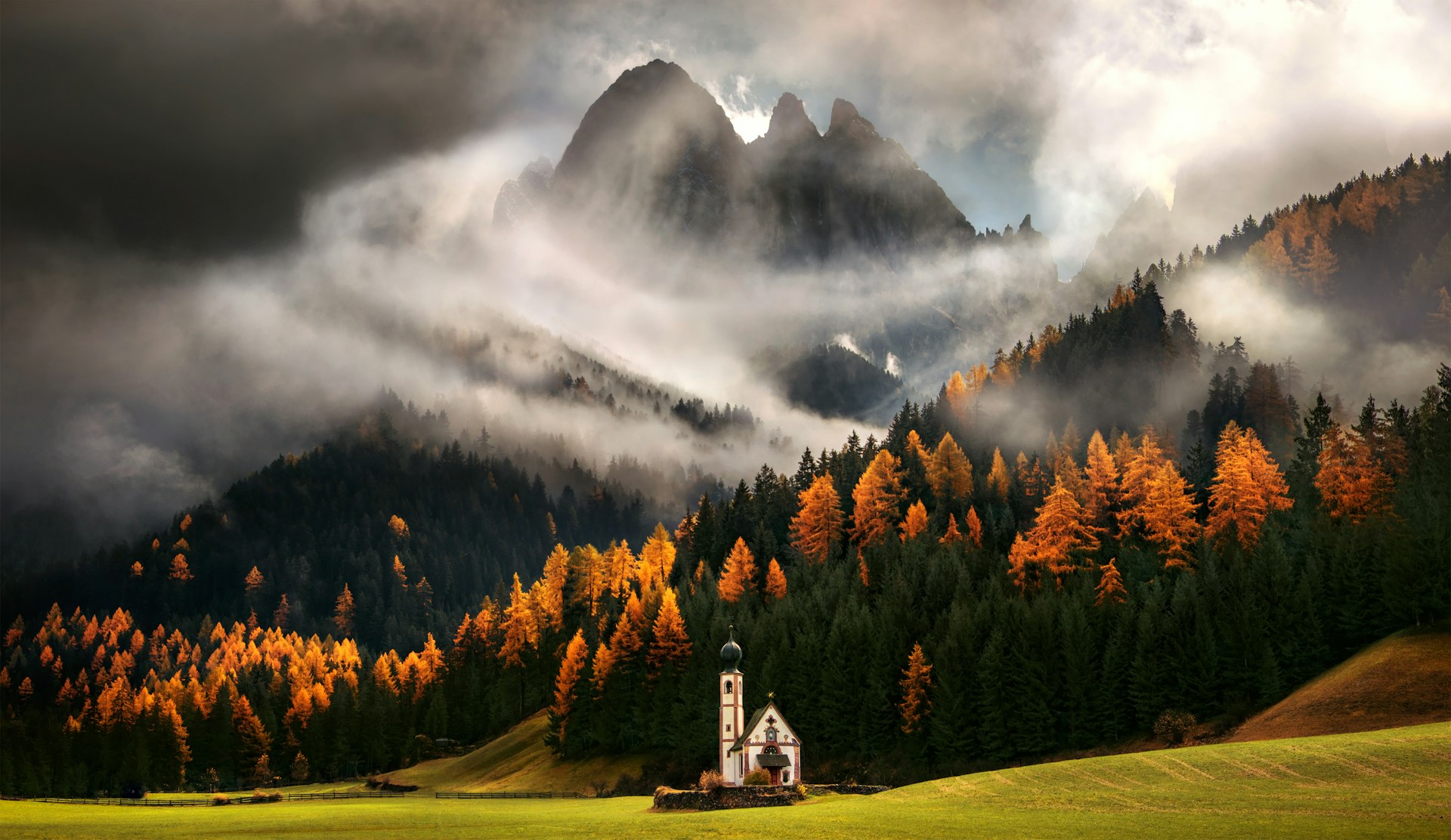 Cloud swirl round craggy Peaks of the dolomites in Italy as forests run down the hillside turning from green into yellows reds and oranges in the autumnal glow with a Russian-style church, Church St. Johann in Ranui, stood at the bottom of the field 