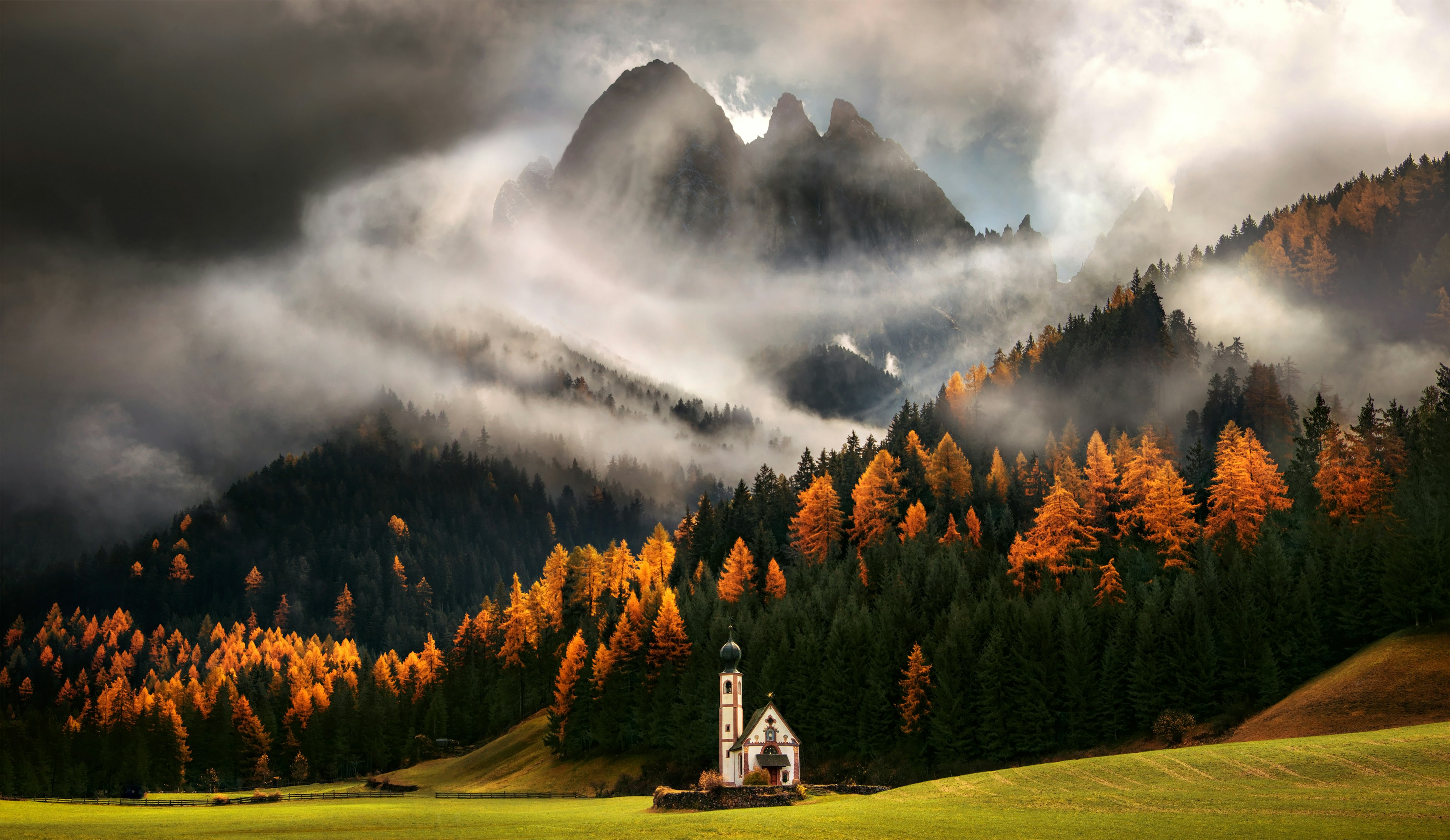 Cloud swirl round craggy Peaks of the dolomites in Italy as forests run down the hillside turning from green into yellows reds and oranges in the autumnal glow with a Russian-style church, Church St. Johann in Ranui, stood at the bottom of the field