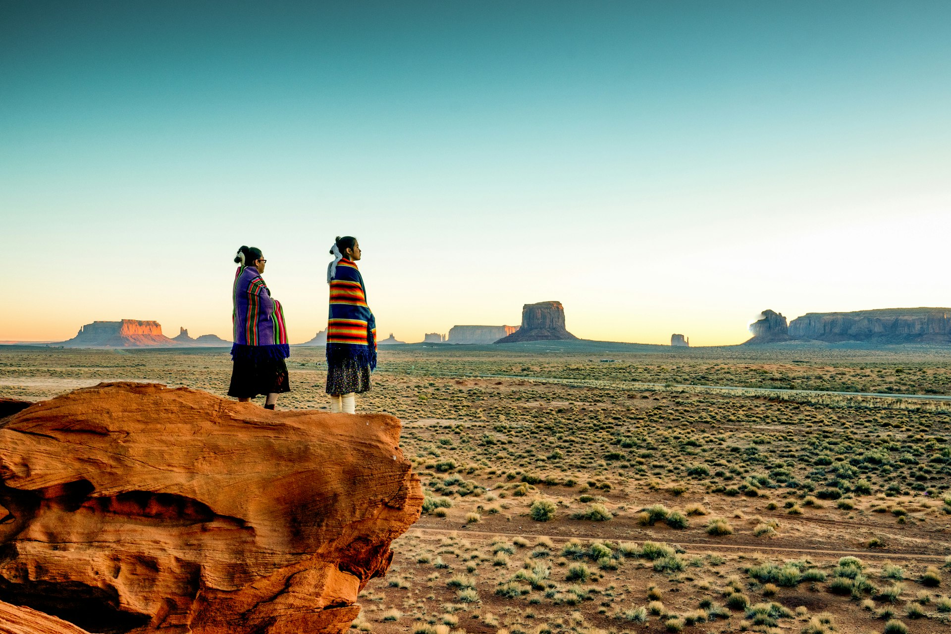 Two sisters wrapped in Navajo blankets stand at the edge of a rock in Monument Valley Tribal Park in the Navajo Nation