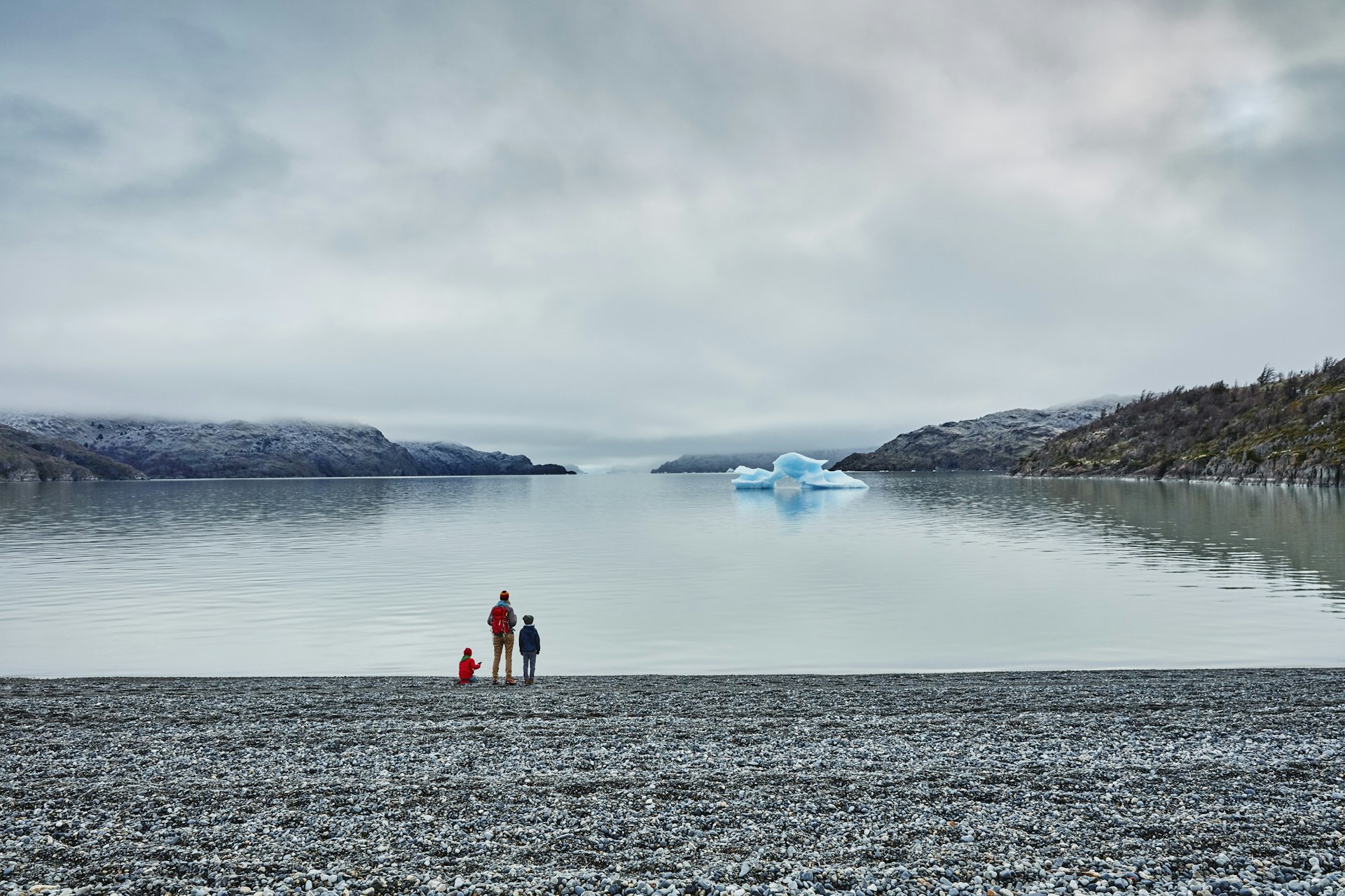 An adult stands with two young children at the edge of a lake looking out towards an iceberg