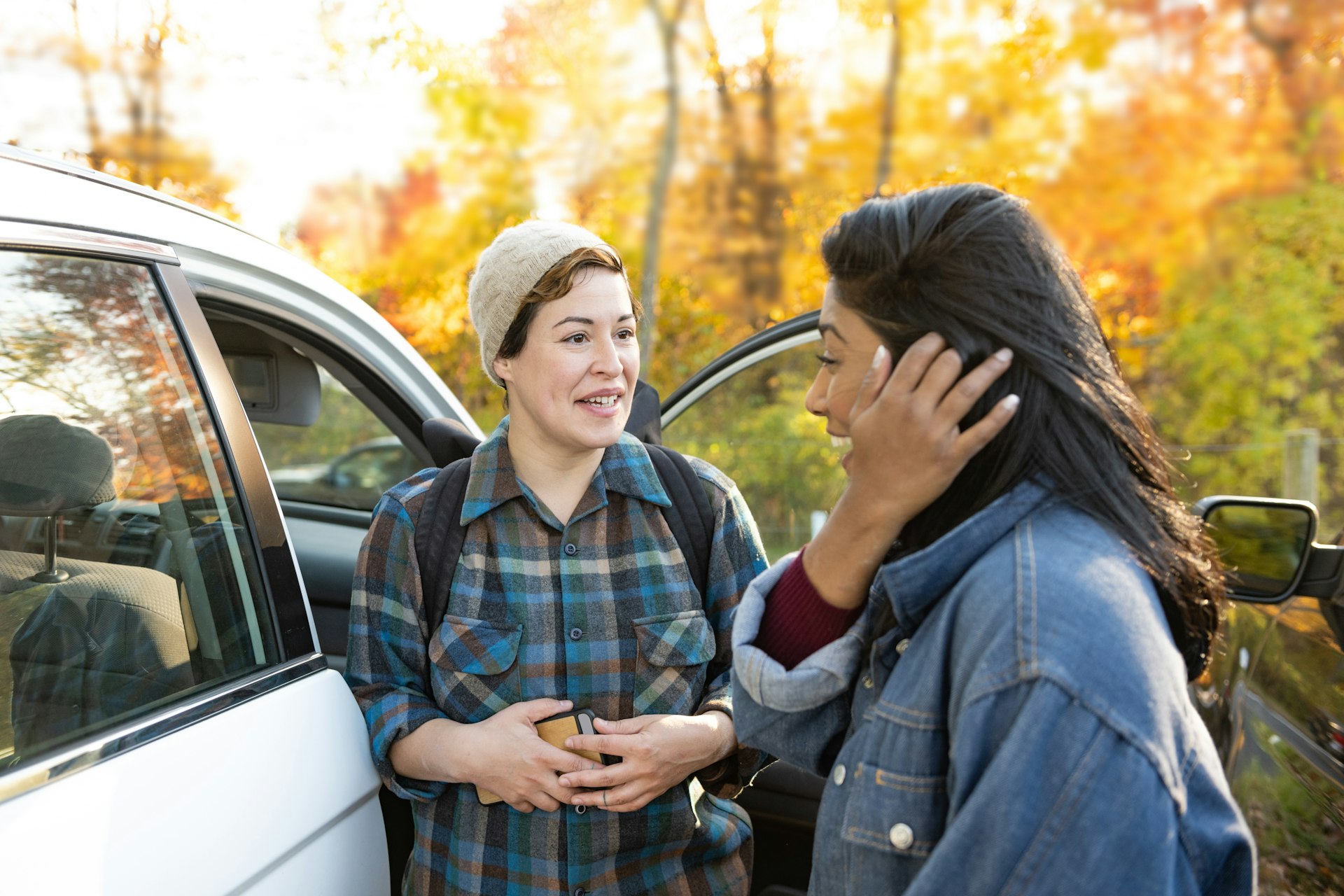 Two adult female friends of different ethnicities greet each other by the car in upstate New York on an autumn day.