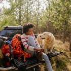 A smiling young woman lacing up her boots on the back of her pick-up truck with her dog next to her.
