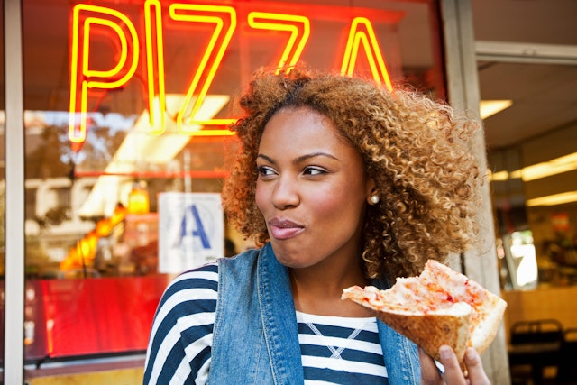 A close up of a woman eating a slice of pizza outside a pizza restaurant in NYC