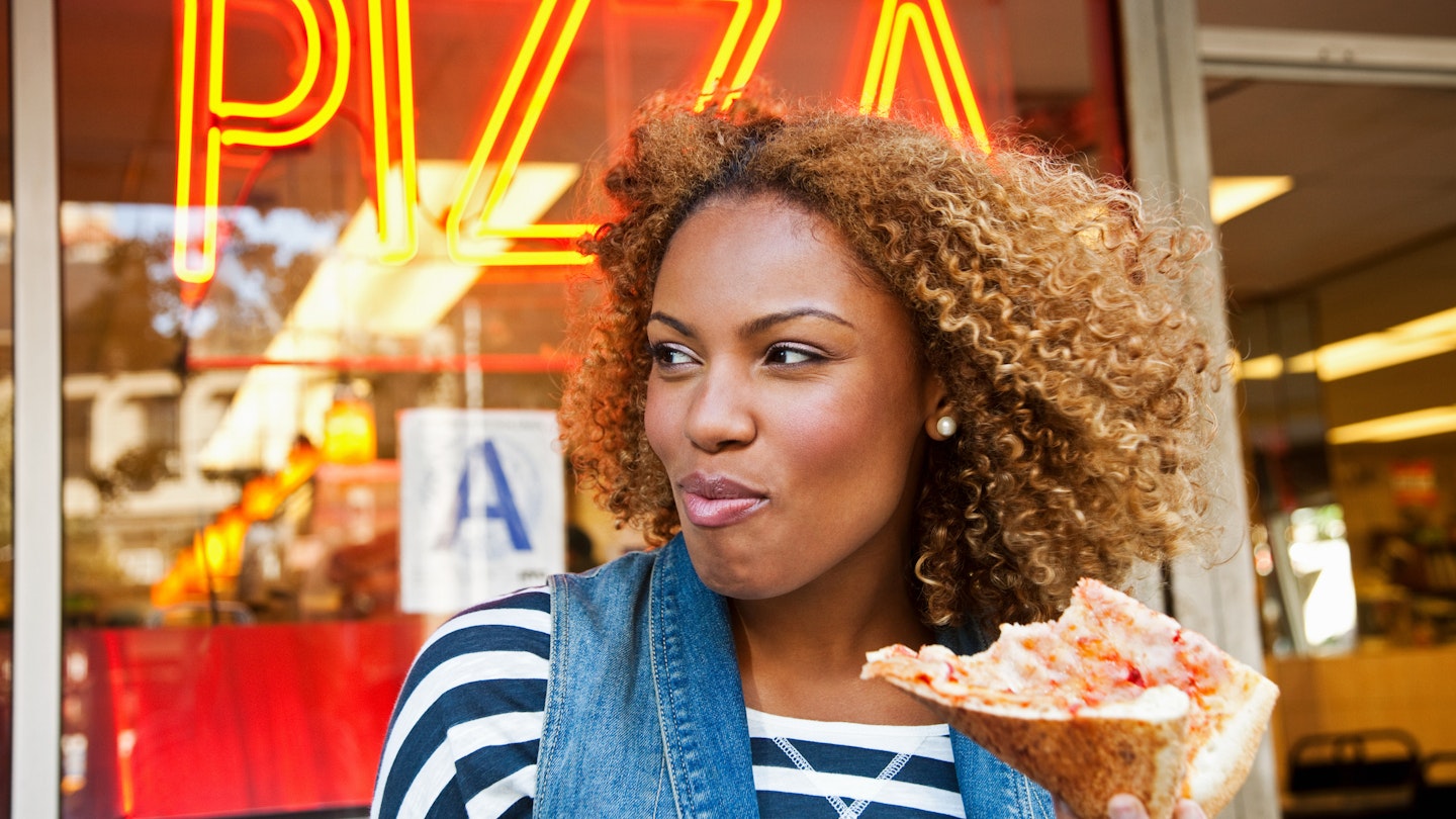 A close up of a black woman eating a slice of pizza outside a pizza restaurant in NYC