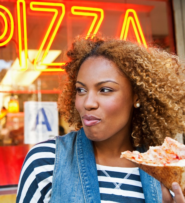A close up of a black woman eating a slice of pizza outside a pizza restaurant in NYC