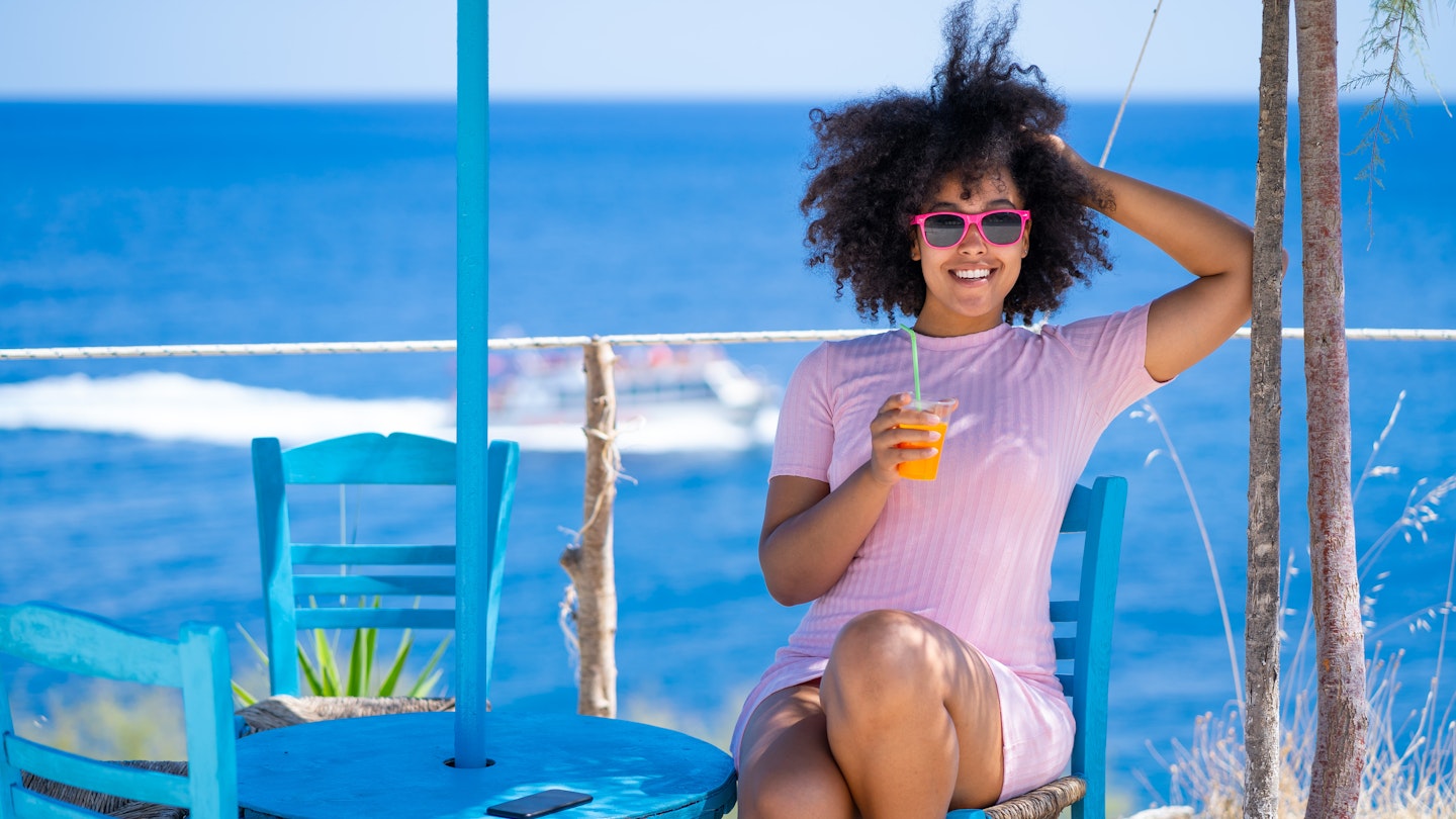 Smiling young woman with sunglasses resting, drinking fruit juice on a beach, sitting on a blue wooden chair
1373066341
A smiling young woman drinking a cocktail at a taverna by the sea in Greece