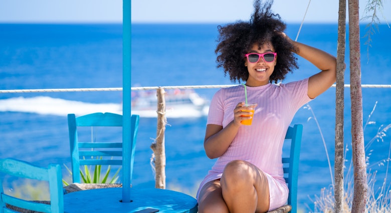 Smiling young woman with sunglasses resting, drinking fruit juice on a beach, sitting on a blue wooden chair
1373066341
A smiling young woman drinking a cocktail at a taverna by the sea in Greece