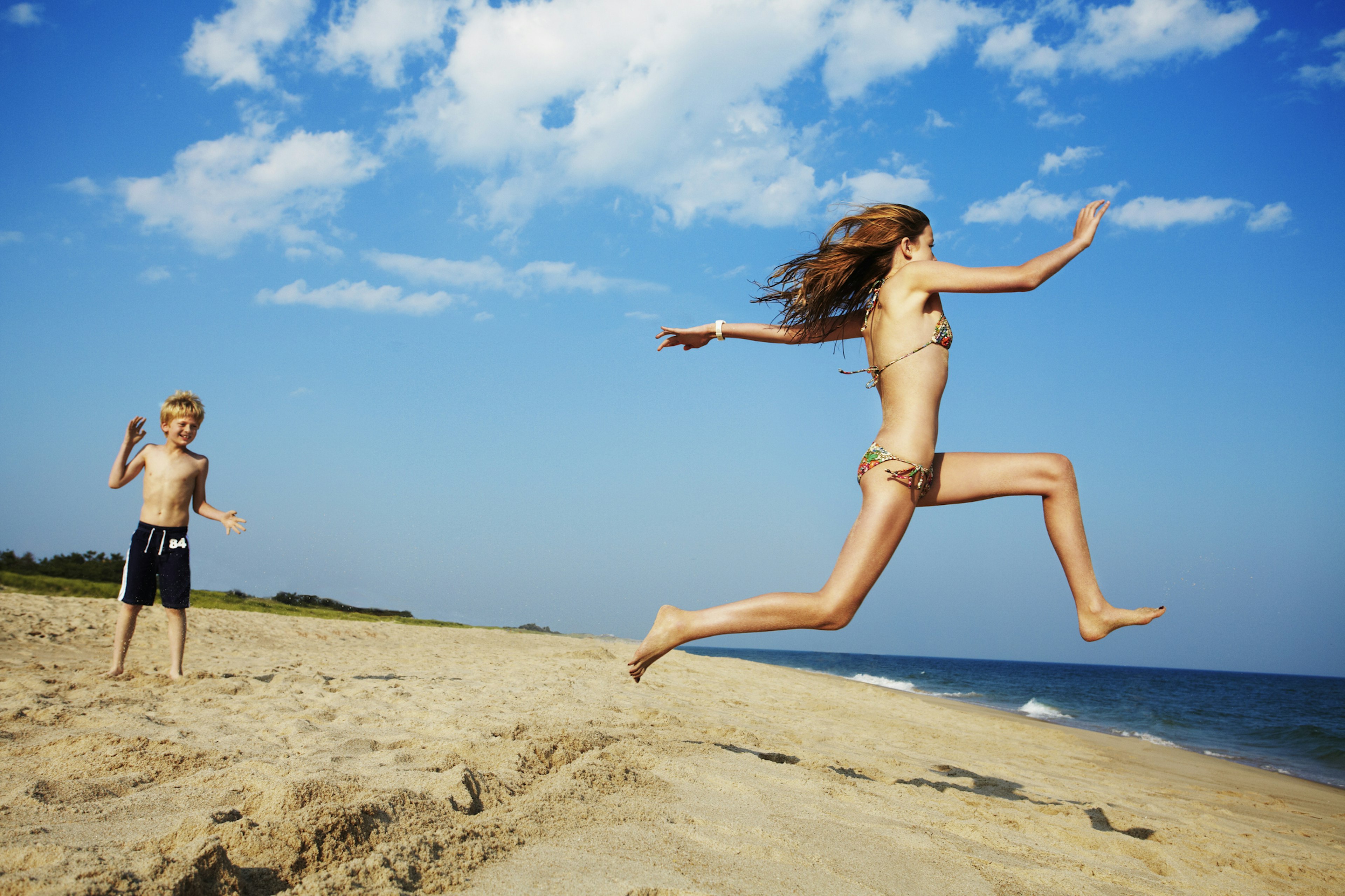 A girl, running along the white sand of the Hamptons in New York State, leaps towards the sea as a younger boy looks on