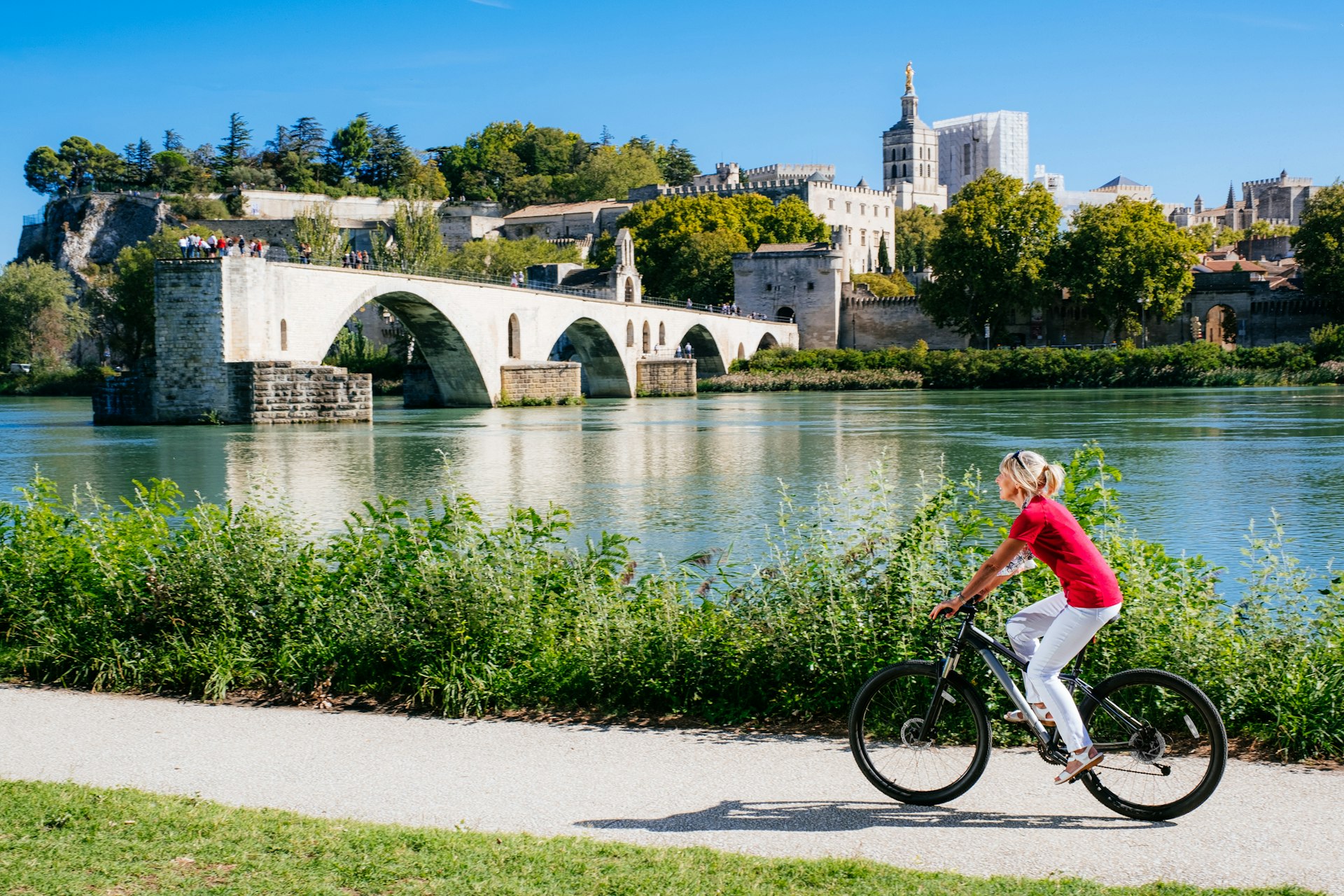 Woman cycling on a path across the river from a medieval bridge that only crosses partway