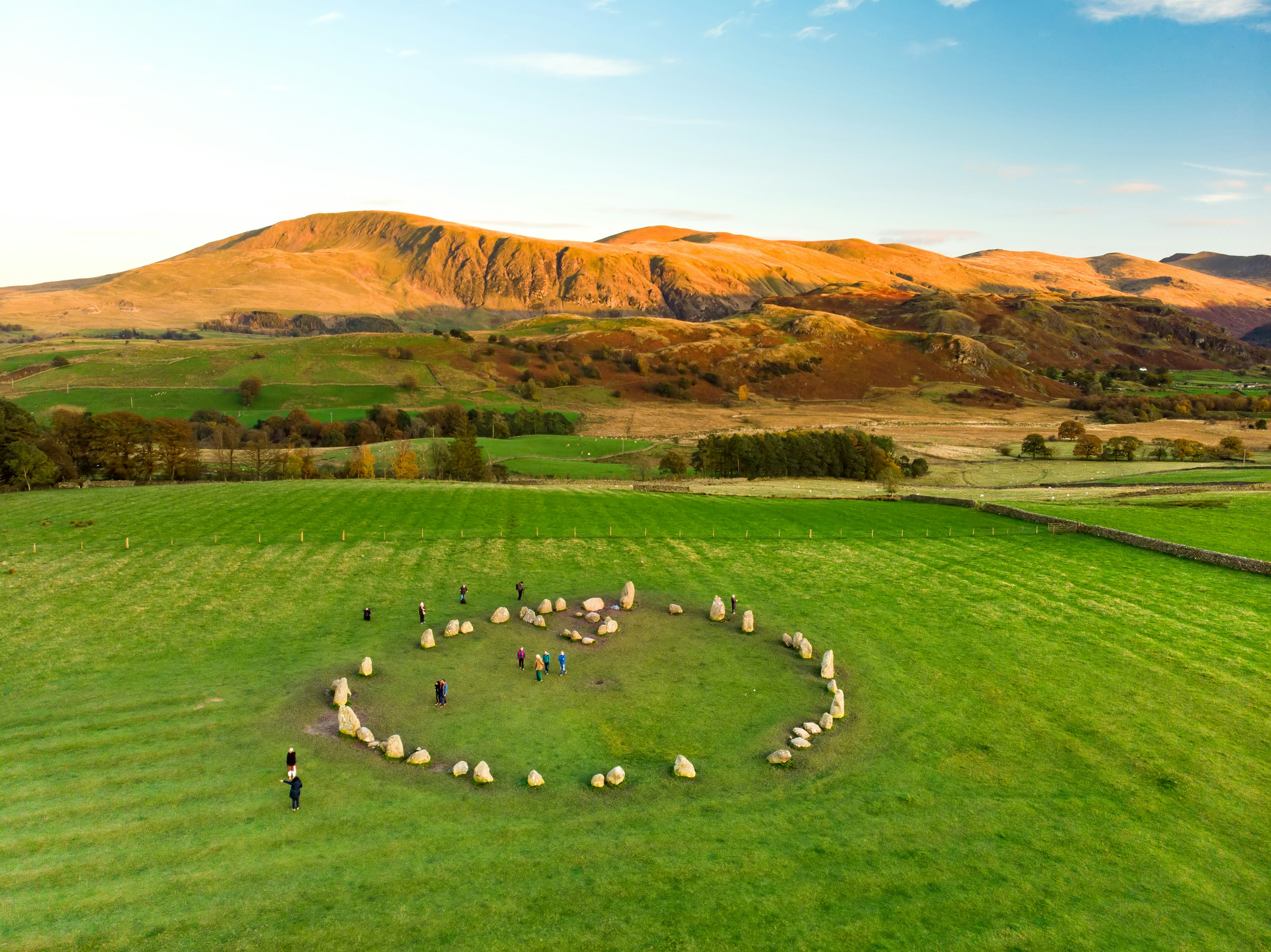 People moving among a circle of standing stones in a rural, hilly setting