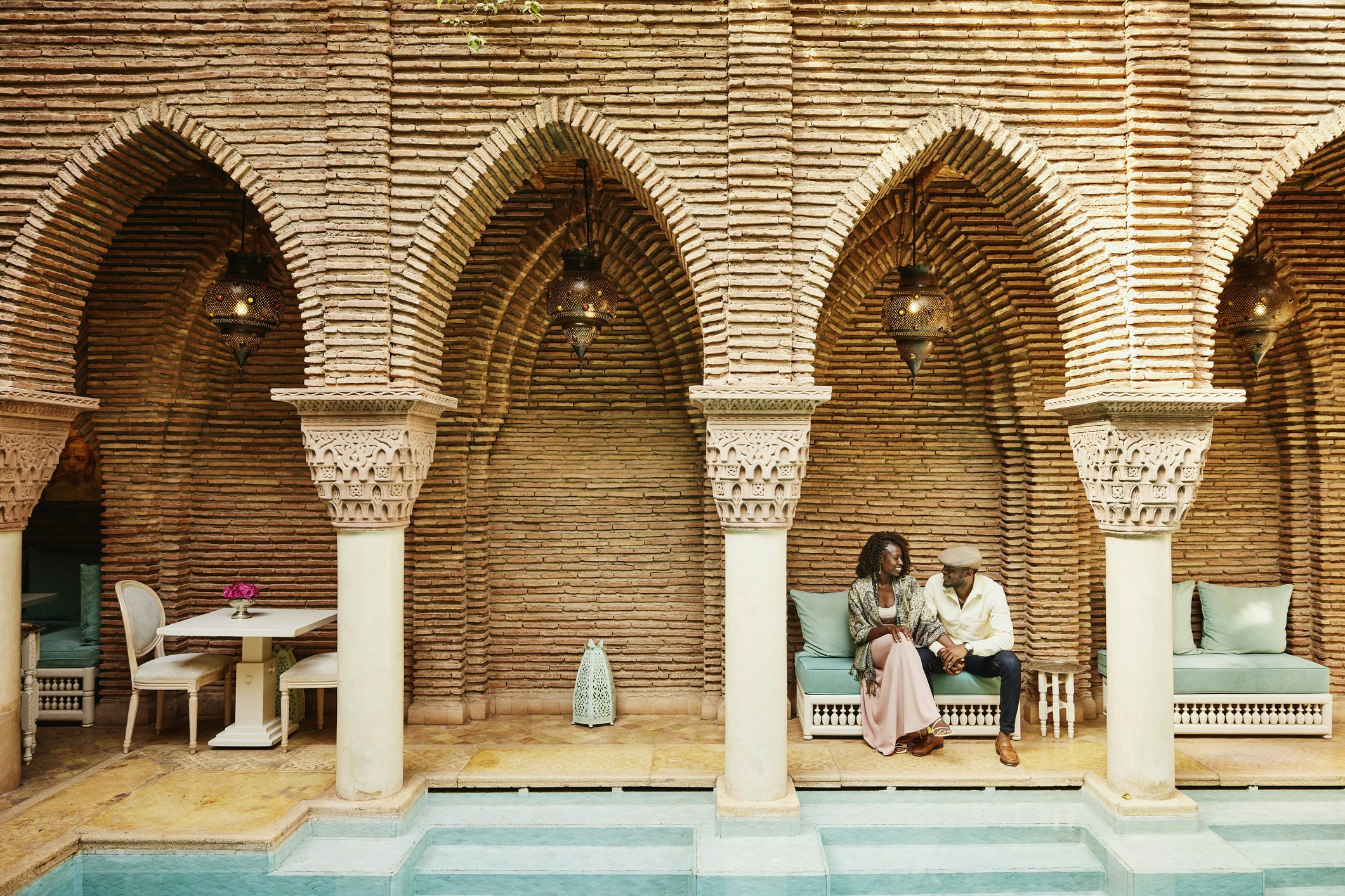 Wide shot of smiling couple holding hands while sitting by pool in courtyard of luxury hotel while on vacation in Marrakesh, Morocco