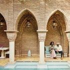 Wide shot of smiling couple holding hands while sitting by pool in courtyard of luxury hotel while on vacation in Marrakesh, Morocco © Thomas Barwick / Getty Images