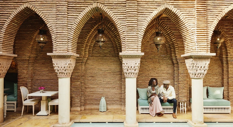 Wide shot of smiling couple holding hands while sitting by pool in courtyard of luxury hotel while on vacation in Marrakesh, Morocco © Thomas Barwick / Getty Images