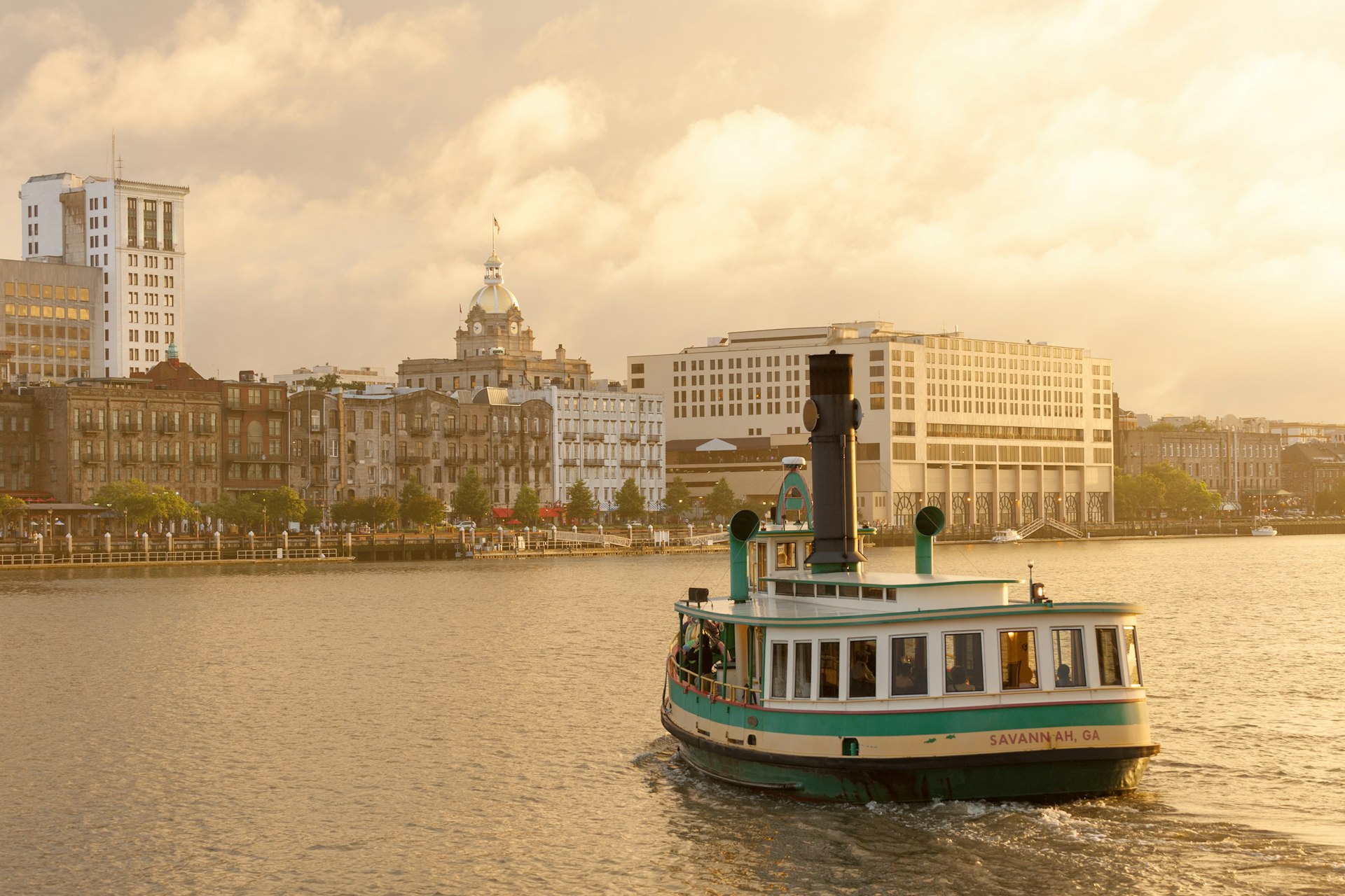 A white and green ferry approaches a historic riverside street from the water in Savannah, Georgia