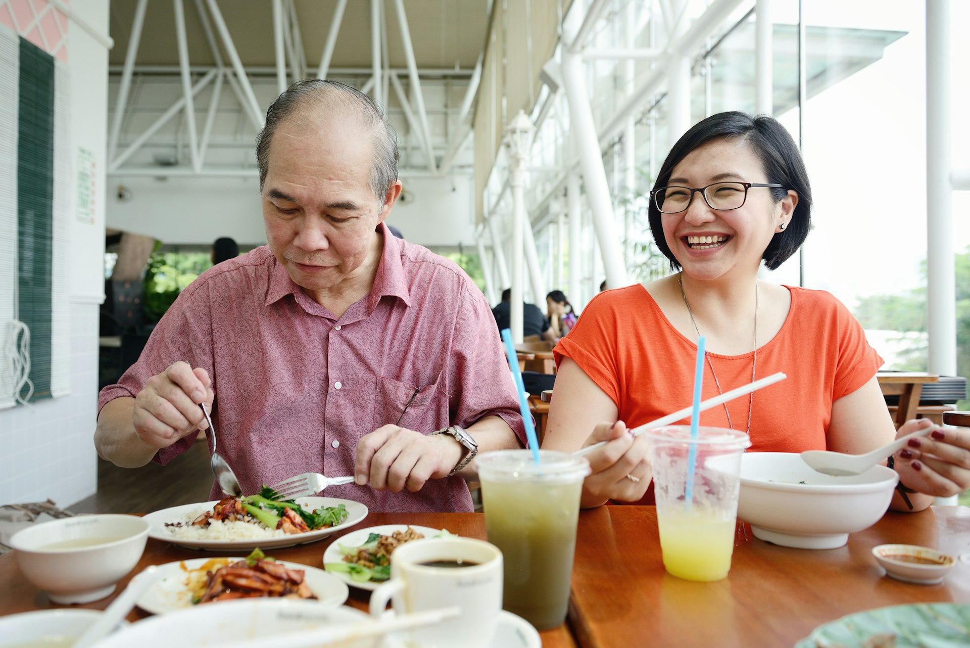 Asian lady and a mature man enjoy their meal. The lady is laughing at something, while the man is concentrating on his food.