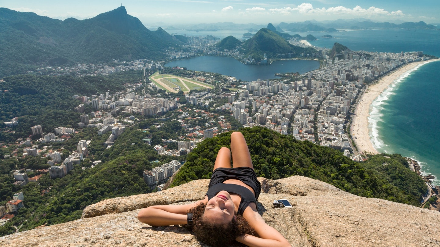 Young mixed race woman in exercise outfit relaxes and sunbathes on the top of Dois Irmaos mountain, with Ipanema beach and lagoon as the backdrop.
560120167