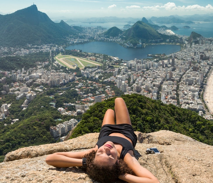 Young mixed race woman in exercise outfit relaxes and sunbathes on the top of Dois Irmaos mountain, with Ipanema beach and lagoon as the backdrop.
560120167