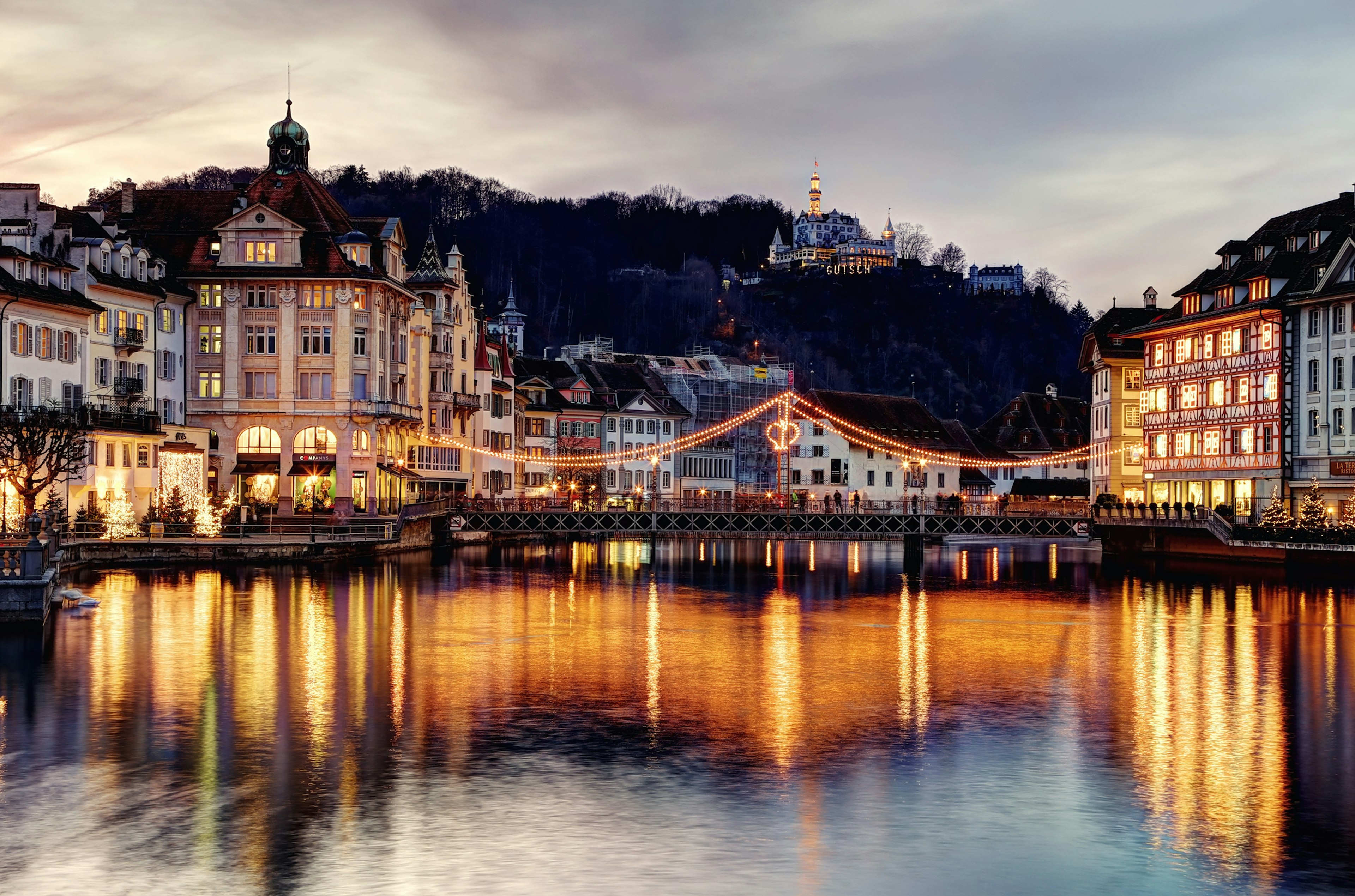 A city based around a river lit up at night. A castle peaks out of woodland at the top of a hill