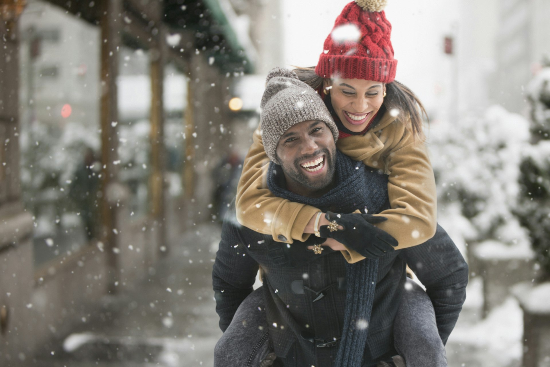 A smiling man gives a smiling woman a piggy back in New York City with as snow fallas around them 