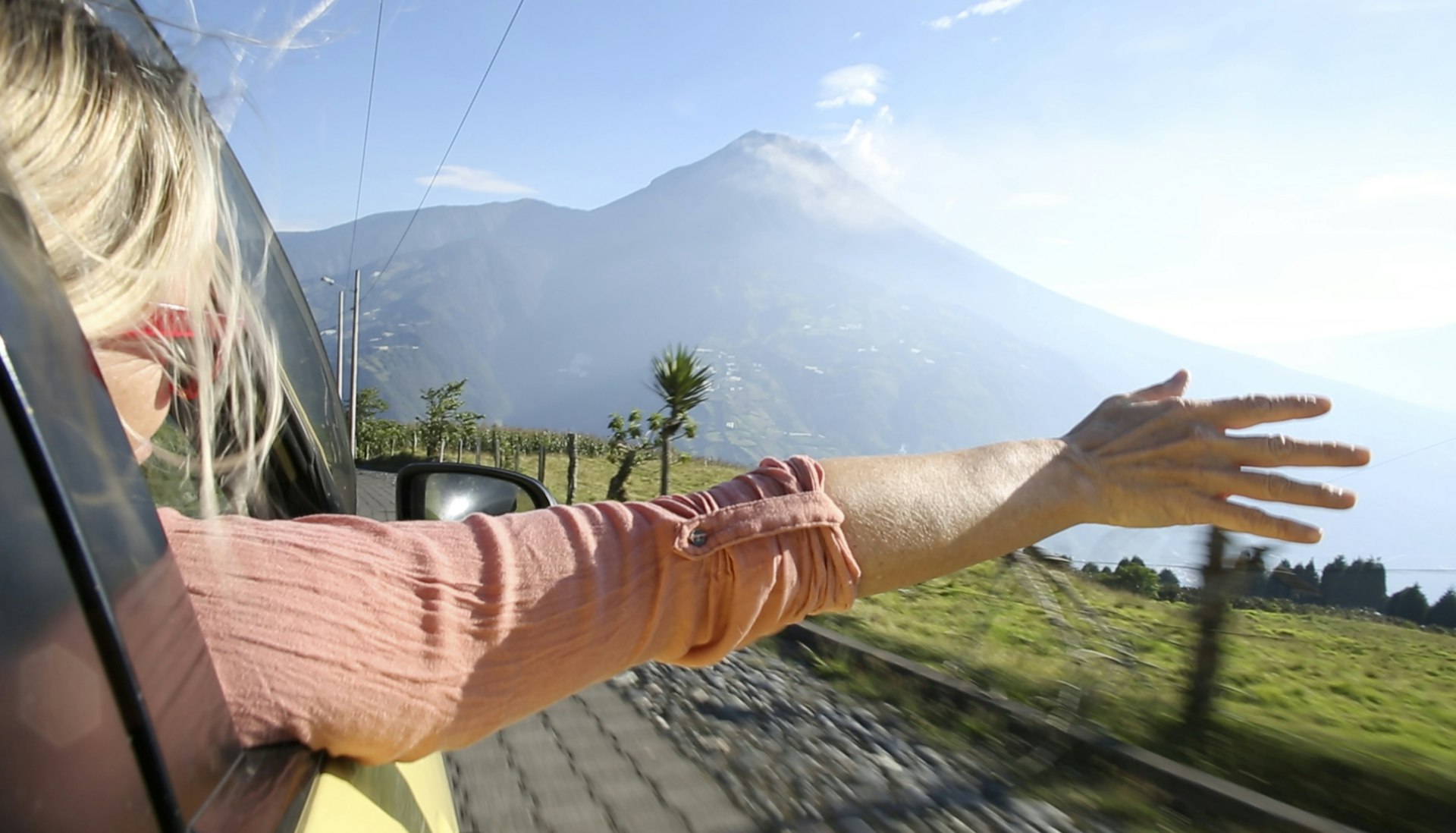 A woman with her arm outstretched from a car window in Ecuador. A mountain is in the background.