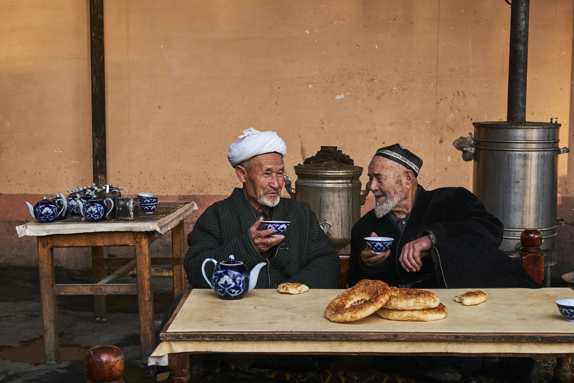 Two men in traditional Uzbek dress chat over tea and bread in a teahouse