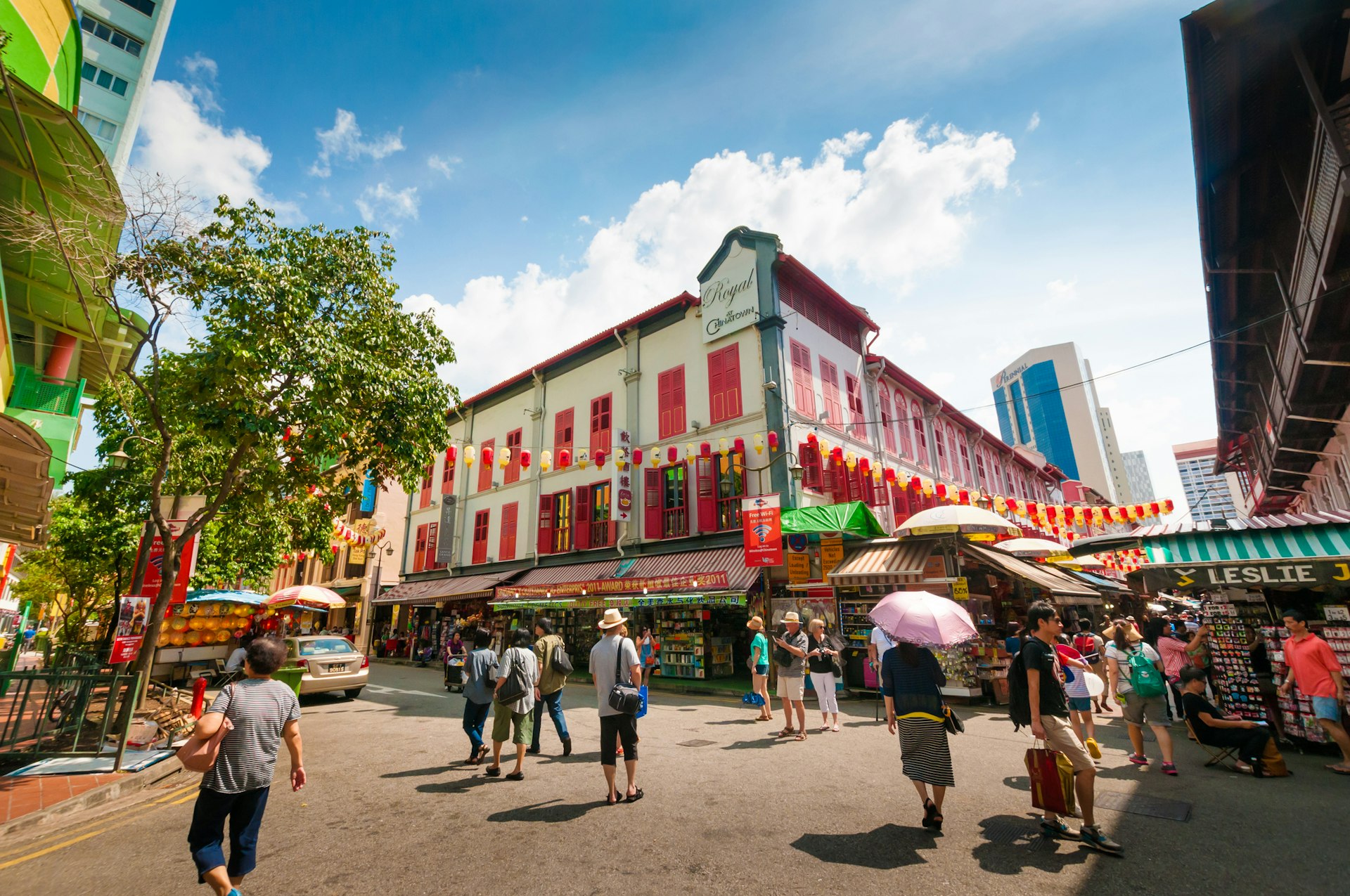 Tourists milling around the street of Chinatown in Singapore as the sun beats down; some carry umbrellas to give them shade