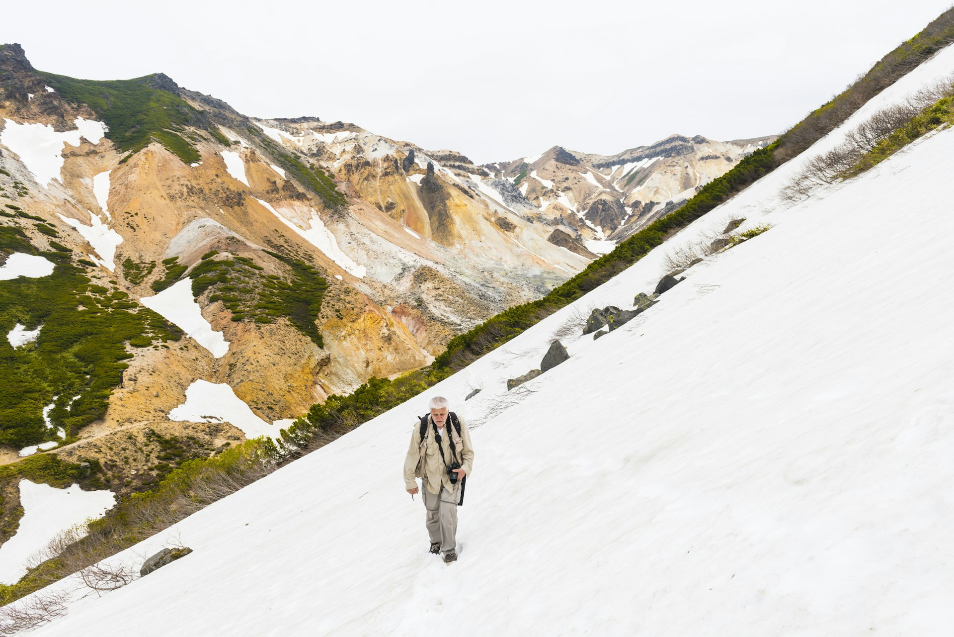 A senior male hiker walks along a snow-covered slope with beige and brown volcanic hills behind him