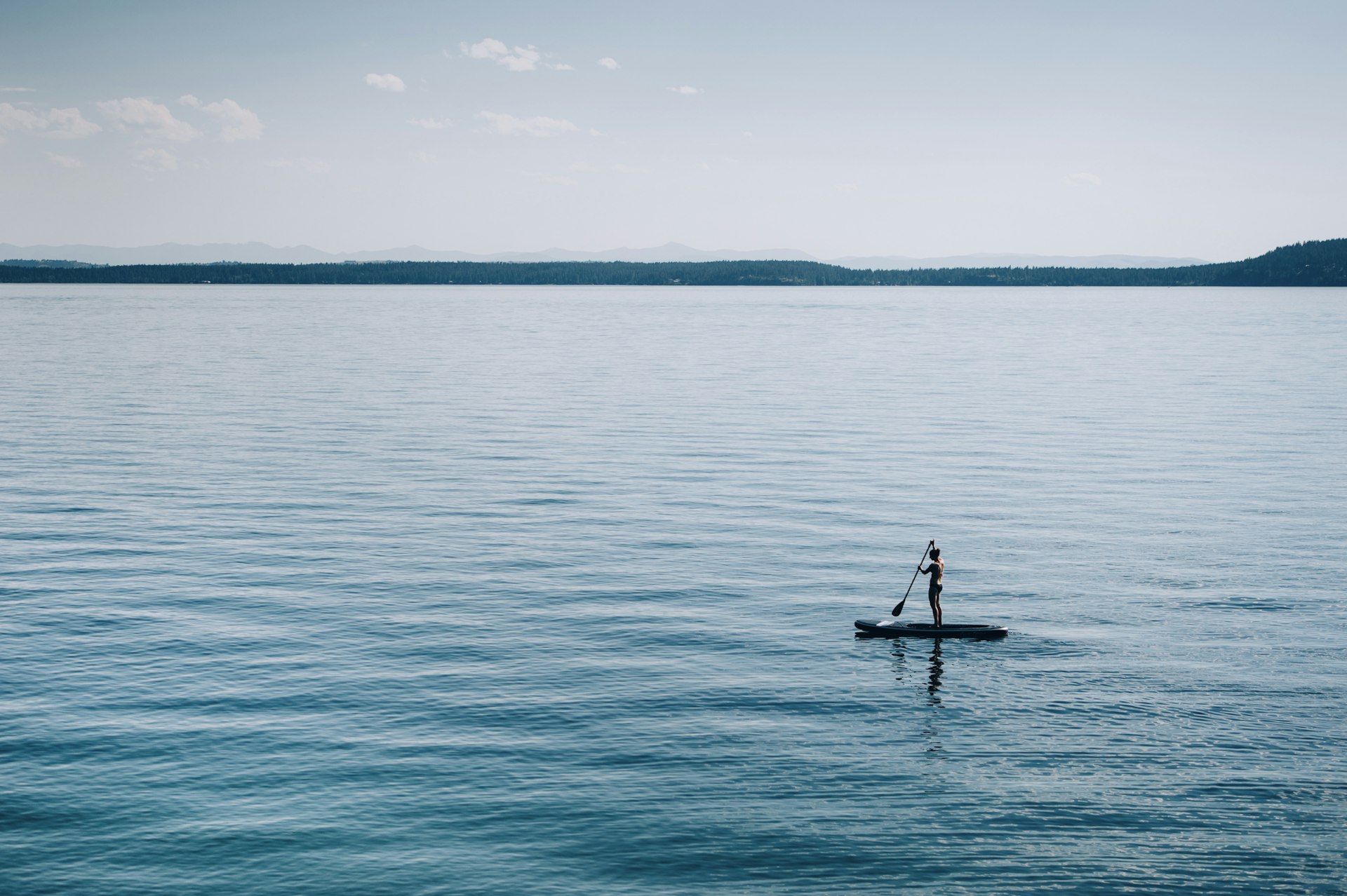 A solo figure stands on a paddleboard on an incredibly still lake surface