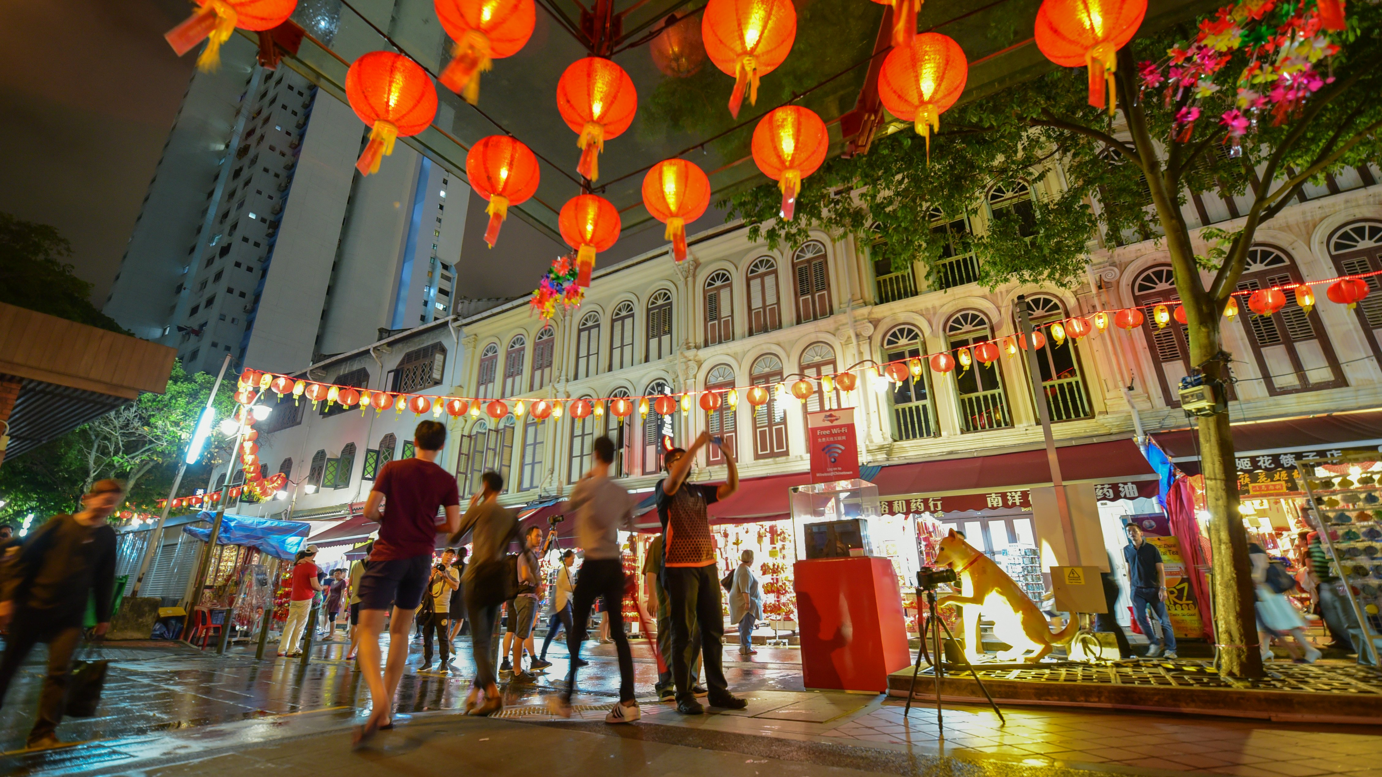 People walking and taking photos of red paper lanterns above a shopping street at night.