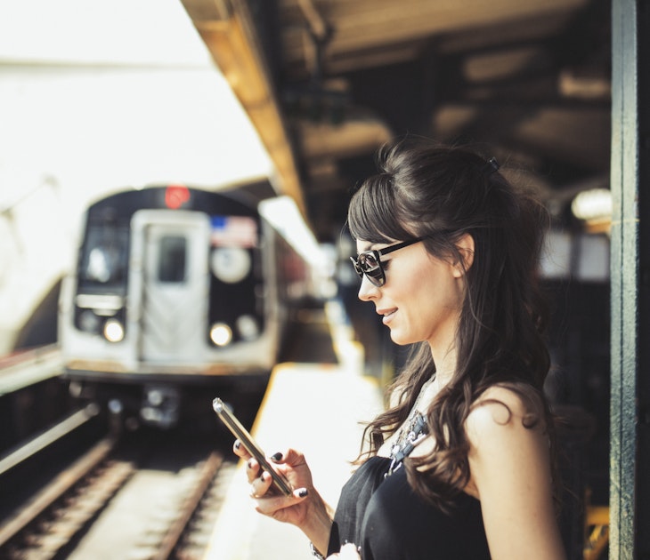 A woman waiting on a platform looking at her phone as a train comes into the station.