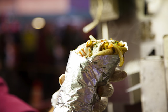 Close-up of food vendor holding a burrito from his food truck, stall on a sidewalk in New York City, USA. The unrecognizable vendor's hand reaches out to show what he has for sale