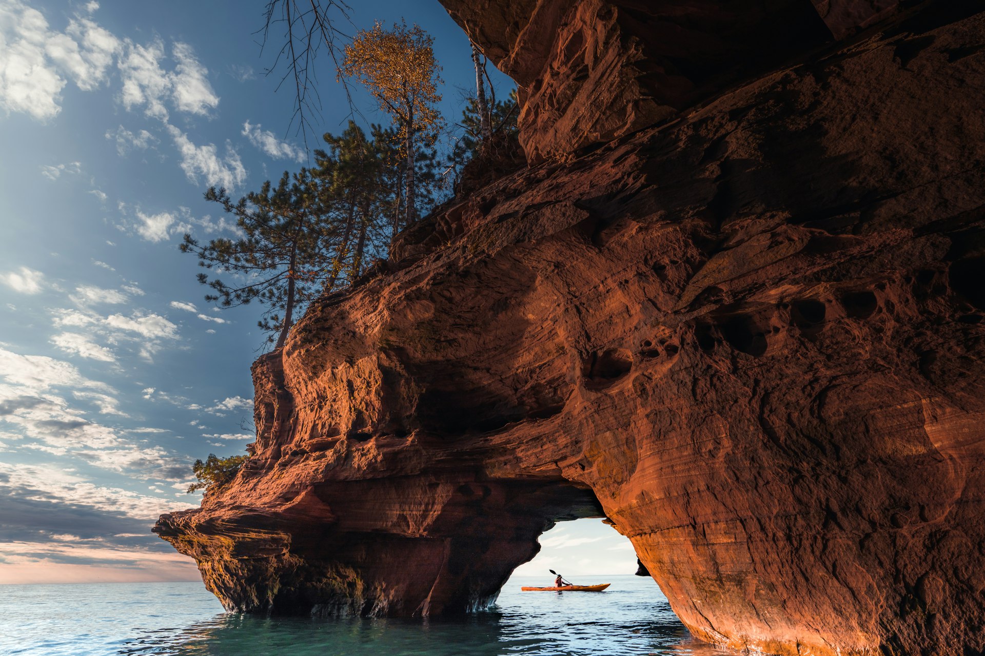 A kayaker viewed through a sea arch of red rock with some trees on top