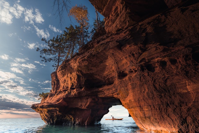 A kayaker viewed through a sea arch of red rock with some trees on top